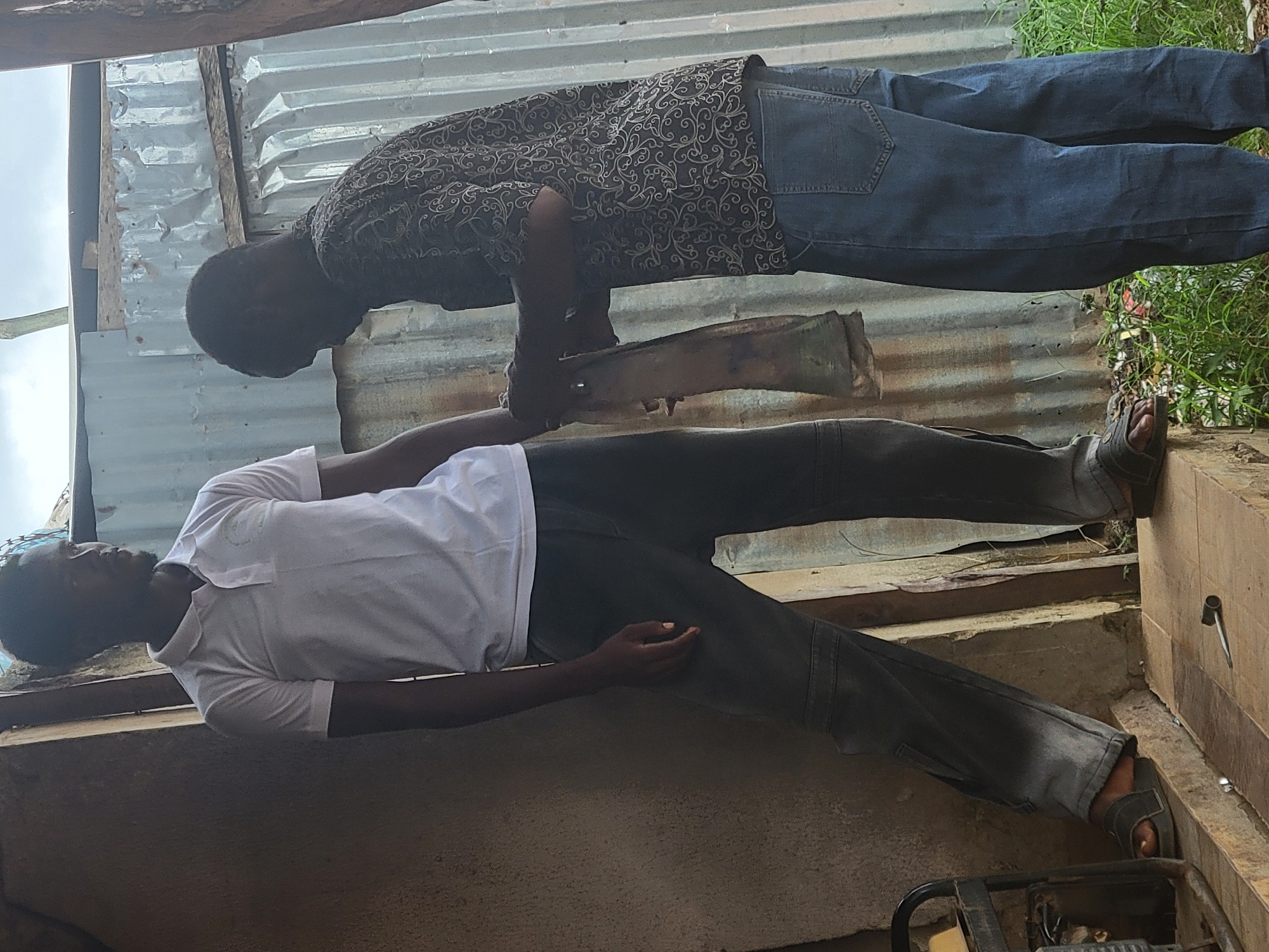 The scene depicts two adult males interacting in an outdoor or semi-outdoor workshop-like setting in the city of Keffi, Nigeria.
The man on the left, with a dark complexion, short dark hair, and a faint beard, wears a white short-sleeved polo shirt with an unidentifiable logo on the right chest, dark grey cargo-style pants with knee paneling, and dark open-toed sandals. He stands mostly upright, facing slightly to his right, with his left foot on a raised, light-colored wooden or concrete platform and his right foot positioned slightly lower. His right hand is near the other man's hands.
The man on the right, also with a dark complexion and head bent down, wears a dark short-sleeved collared shirt with a light-colored, intricate swirling pattern, blue denim jeans, and dark closed-toe footwear. He is bent forward, focused on an object he is holding. Both of his hands are manipulating a light brown, elongated, irregularly shaped object, possibly a piece of animal hide, leather, or rough fabric, which appears to be the subject of their interaction.
The immediate foreground features the raised platform, where a small black and silver tool resembling a socket driver or wrench lies horizontally. To the far left, a black metal frame, potentially part of a generator or equipment stand, is partially visible. The background consists predominantly of weathered, vertically oriented corrugated metal sheets, likely forming a wall or fence. Above the metal sheets, a cloudy sky is visible. The ground where the man on the right stands is uneven and covered with patches of green grass and some scattered debris. To the far right, a rough wooden post is partially visible. The overall lighting suggests natural daylight.