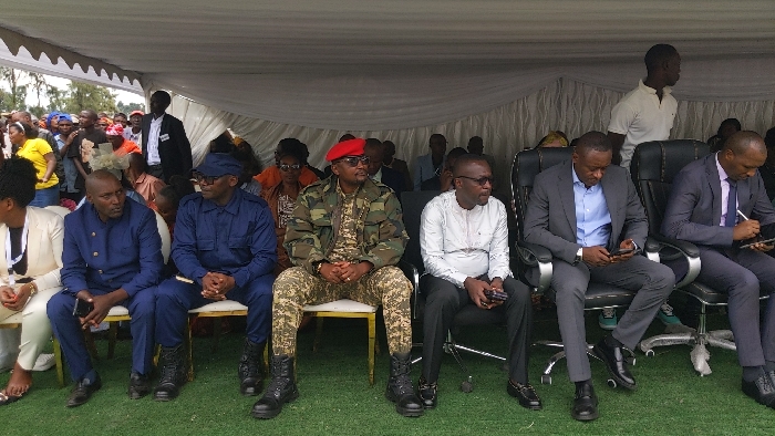 The image shows a group of men sitting in chairs under a white tent. The men are seated in a row, facing forward. The chairs are arranged in a semi-circle. All of the men are wearing formal attire, including suits, jackets, and dress shirts. Some of the men are looking at the camera, while others are looking down or to the side. There are several black chairs in front of the men. The ground is covered in artificial grass. The image was taken in Rukoko, Democratic Republic of the Congo, located at coordinates latitude: -1.624, longitude: 29.258. The scene is a political or public gathering, with officials and possibly other dignitaries present. There is a large crowd of people in the background, suggesting a formal ceremony or meeting.