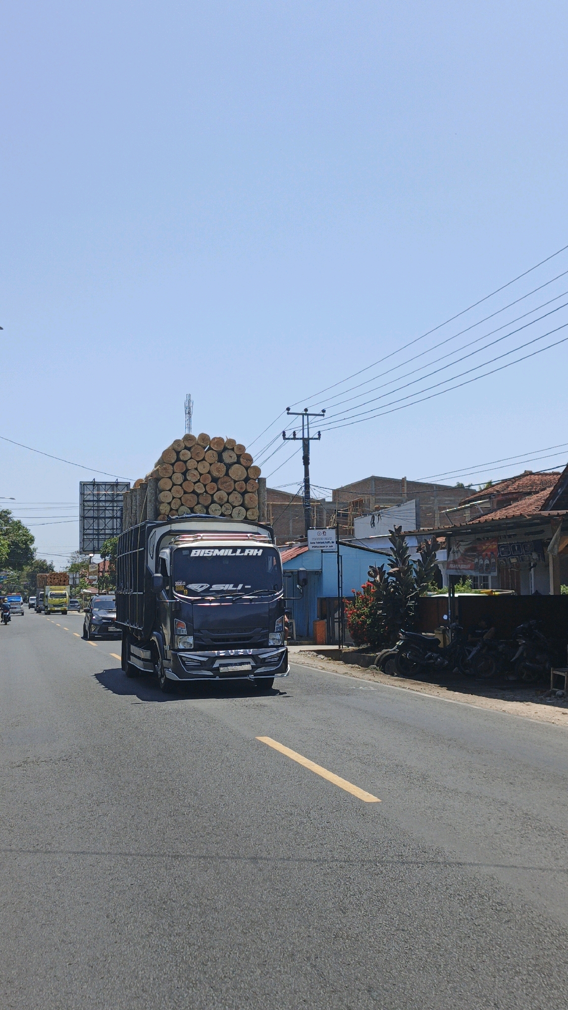 In the city of Desa Wonoharjo, Indonesia, a dark-colored Isuzu truck is driving down a paved road under a clear blue sky. The truck is heavily laden with a large stack of cut logs piled on top of its flatbed. The logs are of varying sizes and are roughly cylindrical in shape. The truck has a prominent "BISMILLAH" sticker above the windshield. The road has a yellow lane divider. In the background, there are buildings, including structures with red tile roofs and some under construction, as well as power lines and a cell tower. To the right of the road, there are several parked motorcycles. Other vehicles, including a yellow truck and cars, are visible further down the road. The overall scene depicts a sunny day in a semi-urban or rural environment.