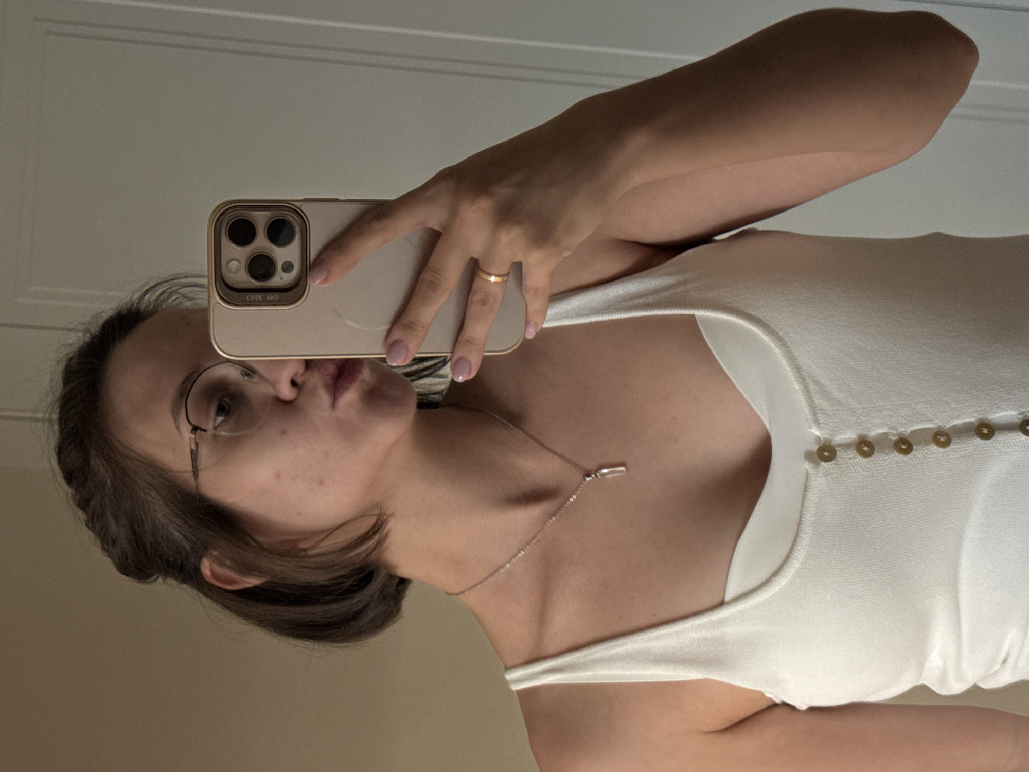 A young woman with brown hair styled in a braid and bob, wearing glasses and a white tank top, takes a mirror selfie. She is wearing a silver necklace with a clear pendant and a gold ring on her right hand. Her nails are painted a light pink. She is holding a gold-colored smartphone with a multi-lens camera. The background shows a wall and a door. The image does not contain any elements that can be used to determine a specific location within the city of Pervomaysky, Russia.