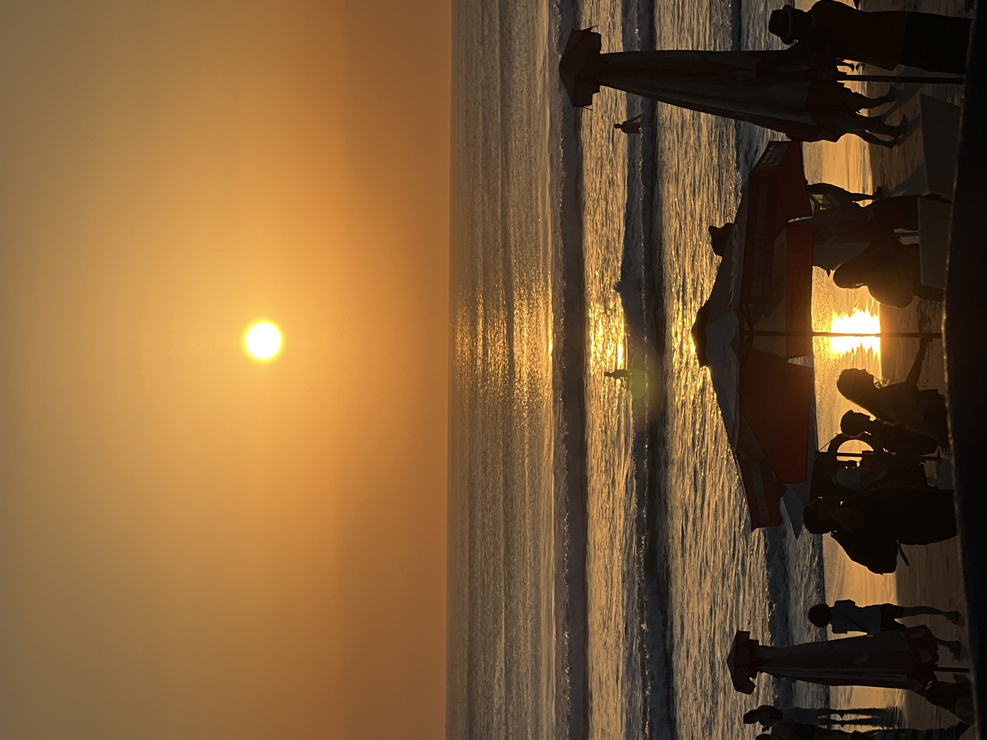 The image depicts a vibrant sunset over the ocean at Kuta Beach, Indonesia. The sun, a bright orb of orange, sits low on the horizon, casting a golden glow across the sky and the water. Gentle waves roll towards the shore, their crests illuminated by the setting sun. In the foreground, silhouetted figures of people gather on the beach, some standing and some walking. Beach umbrellas, some partially furled, dot the sandy expanse, adding to the scene's relaxed atmosphere. The overall impression is one of tranquility and the end of a beautiful day in this popular Indonesian tourist destination.
