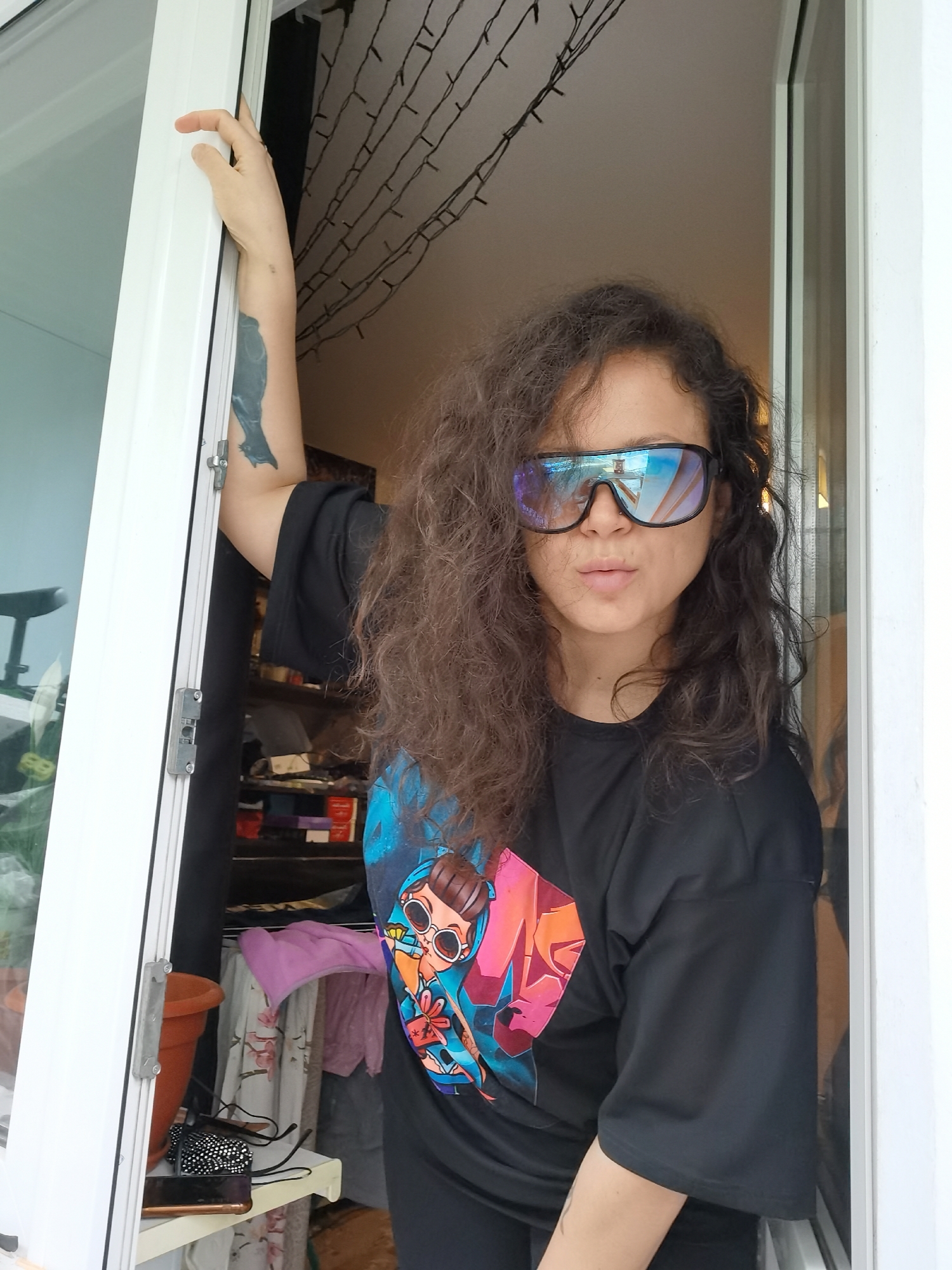 A woman with long, voluminous dark brown curly hair is pictured leaning through a white UPVC door or window frame in Chaikovsky, Russia. She looks directly at the camera, her mouth slightly pursed. She wears large, reflective, rectangular sunglasses that obscure her eyes, and a small stud nose piercing is visible.
She is dressed in a loose, black short-sleeved t-shirt featuring a vibrant, colorful graphic print on the front. The print depicts a stylized female character with large sunglasses, holding what appears to be a wrapped gift or a beverage. Her right arm is raised and bent, with her hand grasping the top of the door/window frame. A dark tattoo, possibly of a bird or similar design, is visible on the upper part of her right arm.
The background behind her right side (viewer's left) appears to be a brighter area, potentially an outdoor space or another room. To her left side (viewer's right), an indoor setting is visible, cluttered with various objects. This area includes shelves with items, folded laundry (a pink towel and patterned fabrics), a terracotta plant pot, a black textured bag, and a smartphone resting on a white ledge. Above, strung fairy lights are draped against a light-colored ceiling. The lighting suggests it is daytime. No explicit text is visible.