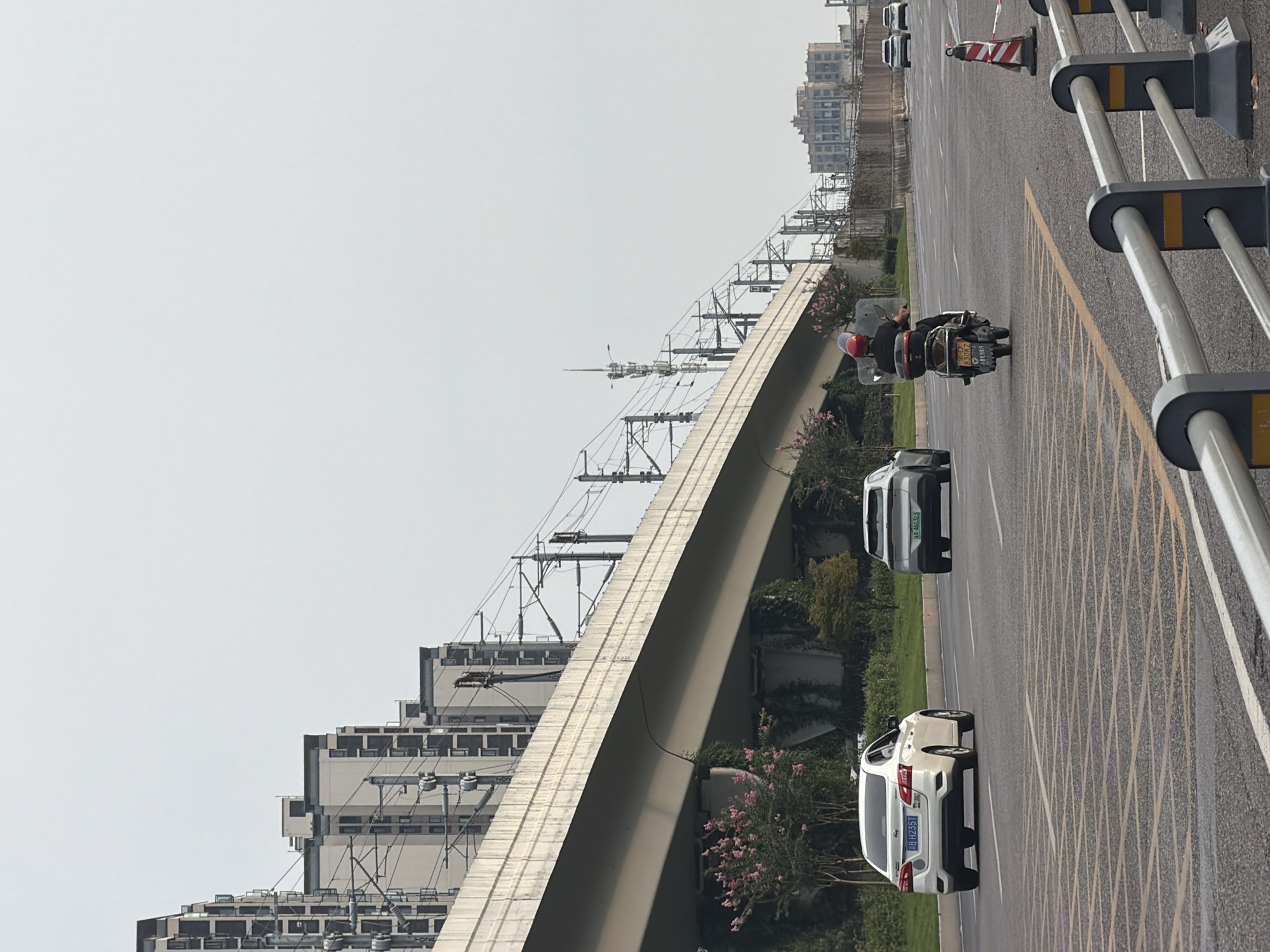 The image depicts a street scene in the city of Jiangjin, China, with an elevated railway line running diagonally across the frame. In the foreground, a paved road displays yellow grid markings for traffic control. A white sedan is visible on the right lane, and a motorcycle with a rider wearing a red helmet is in the center lane, moving away from the viewer. The rider's face is obscured by a shield. Further down the road, two white SUVs are present. The elevated railway structure is supported by concrete pillars, with lush green vegetation and pink flowering bushes growing beneath it. In the background, a series of modern high-rise apartment buildings line the left side of the image, and a larger building complex is situated on the right. The sky is overcast and a pale gray.