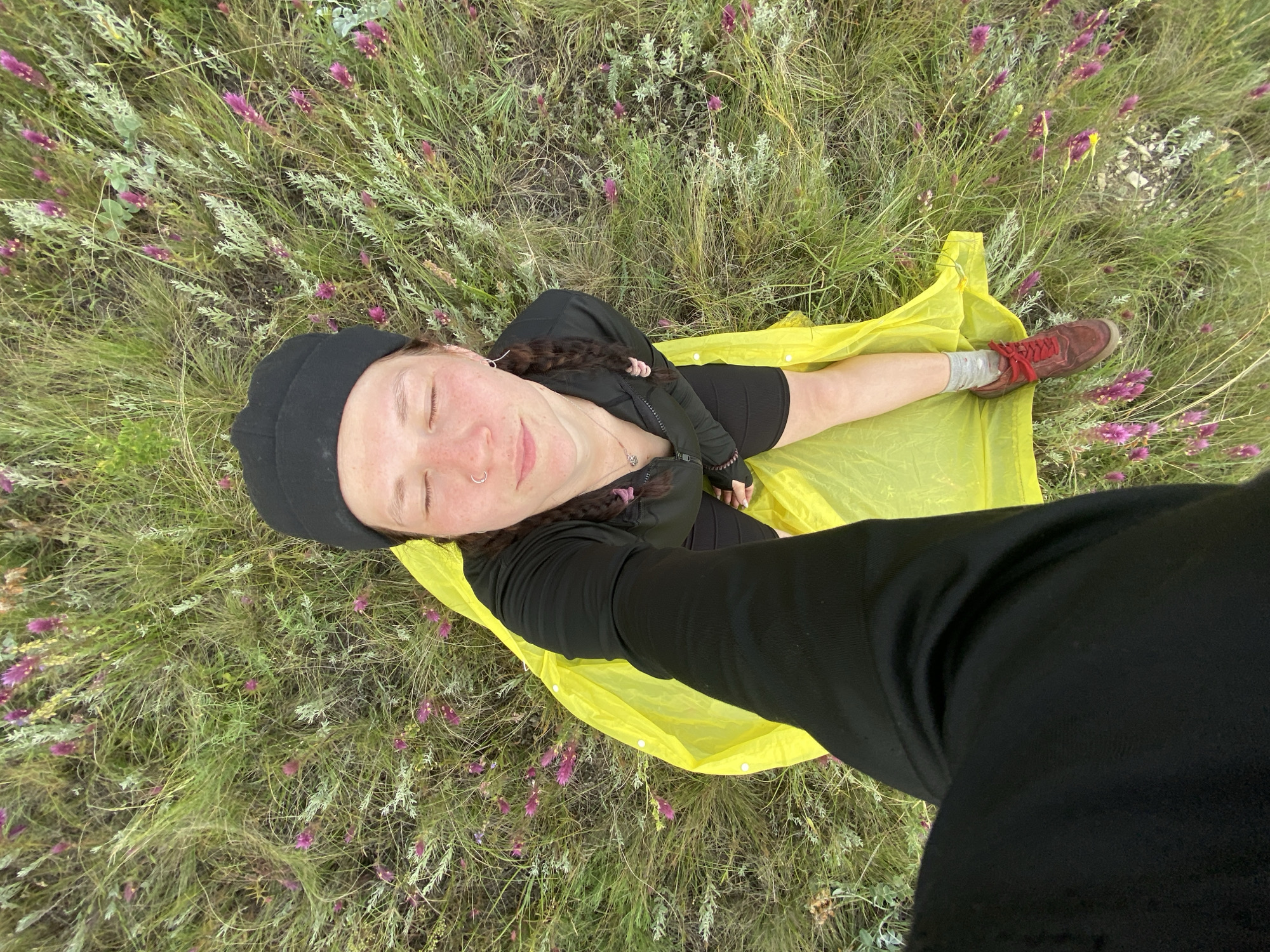 A high-angle, waist-up selfie shows a person with closed eyes, wearing a black beanie, black long-sleeved shirt, and black shorts, lying on a bright yellow ground cover in a grassy field with scattered purple flowers. Their right arm is extended, holding the camera. A person in black pants is partially visible in the lower left corner, suggesting they are taking the photo. The location is identified as Ziyanchurino, Russia.