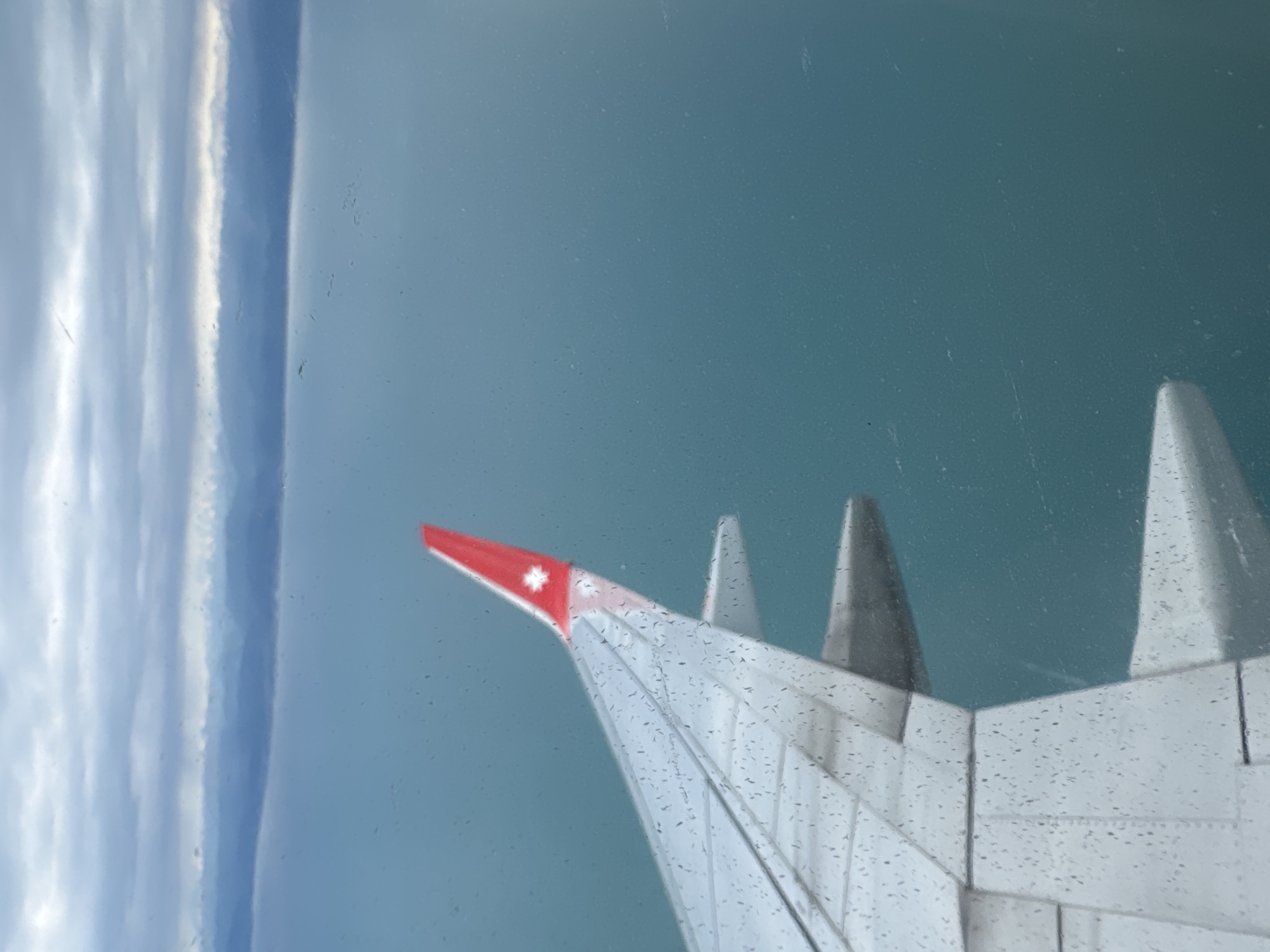 The image shows an airplane wing with a red winglet bearing a white cross design. The wing is mostly white with gray accents. The background consists of a light blue, seemingly endless body of water and a strip of sky with dense, low-hanging clouds. The view is presumably taken from inside the airplane. I cannot determine the location as Sochi, Russia from this image alone.