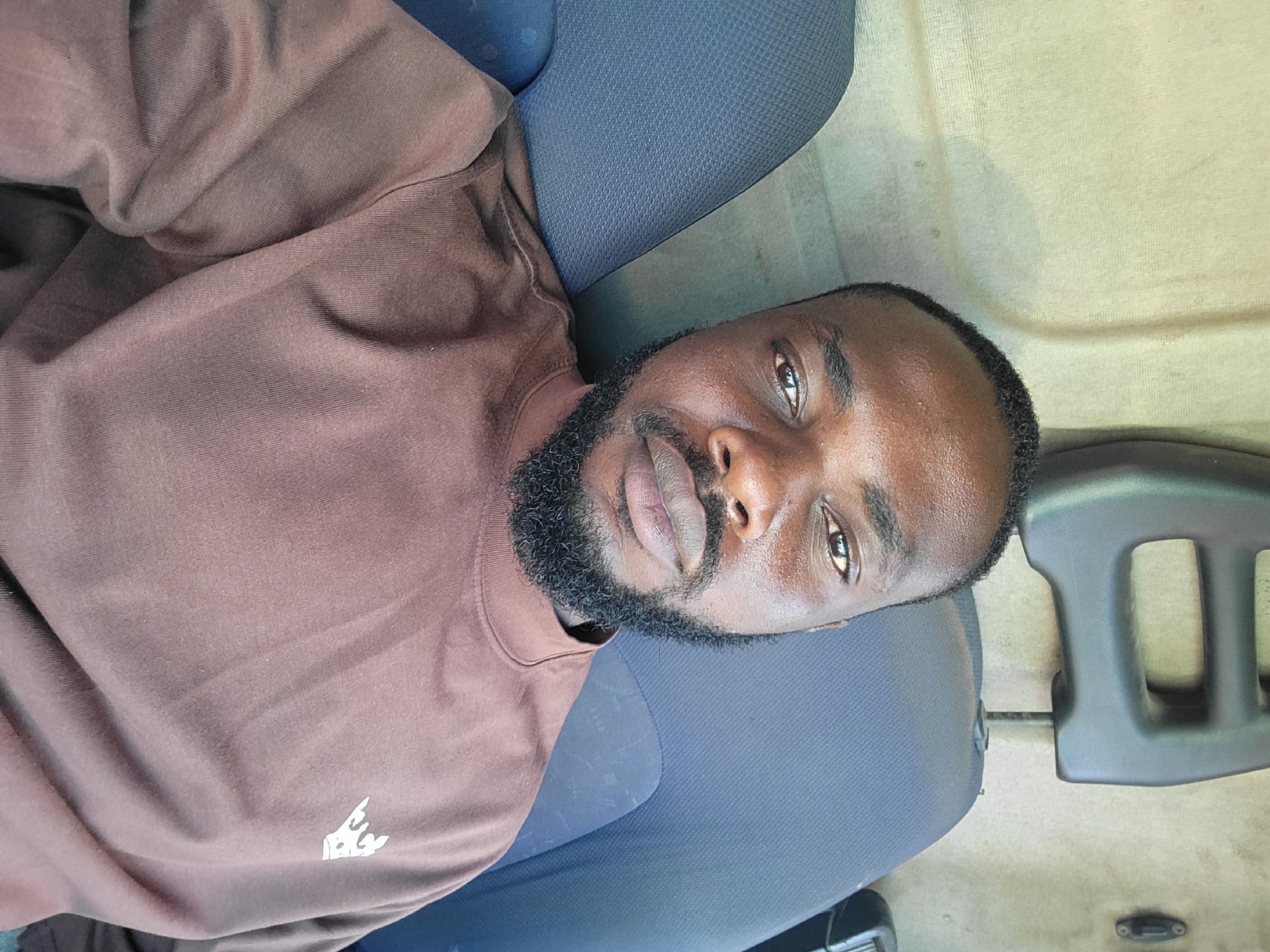 A mid-shot depicts a Black male with a trimmed beard, wearing a brown sweatshirt, laying on a blue fabric surface. The scene appears to be inside a vehicle, considering the interior parts and upholstery. The location within the city of Jos, Nigeria is not discernible from the visual data. The lighting appears to be natural. There are no evident actions or interactions taking place.