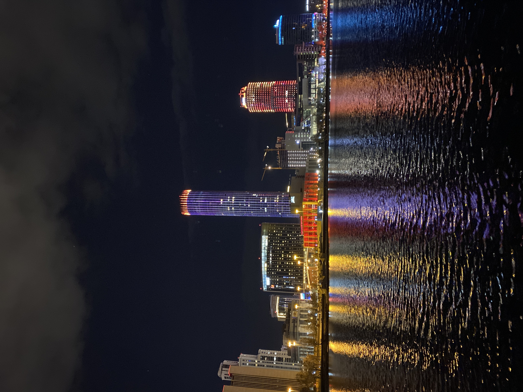 The image shows a nighttime view of a cityscape reflected in a body of water. The cityscape features several illuminated buildings, including a tall skyscraper with purple and orange lights, a round building with red and yellow lights, and several other buildings with varying designs and light schemes. The reflections in the water mimic the colors and patterns of the building lights. Some clouds are visible in the sky. The location is likely the Iset River in Yekaterinburg, Russia, reflecting the Yekaterinburg City skyscrapers.