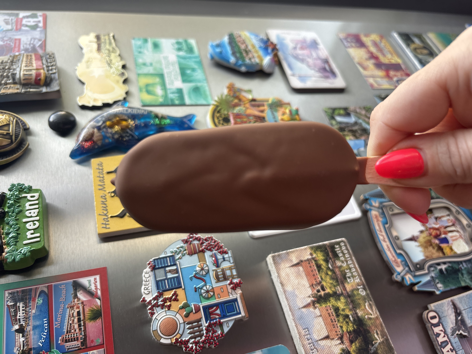 The image depicts a hand holding a chocolate-covered popsicle in front of a refrigerator door covered in magnets. The magnets feature various locations, including Ireland, Greece, and Bulgaria. Notably, one magnet mentions "Duni, Bulgarien" and "Marina-Beach" along with "Pelican", suggesting it might be a coastal resort area in Bulgaria. Another magnet is labeled "The Castle of Rabadinovo". Additionally, there is a magnet mentioning "Hakuna Matata" and another "Halkidiki". The background is a metallic surface, likely the refrigerator door. The hand is manicured with bright red nail polish.