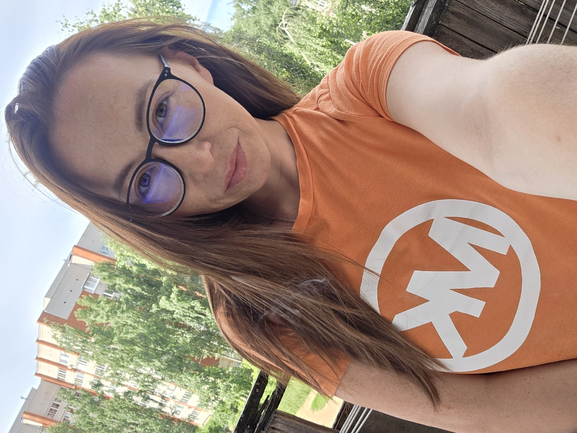 A medium shot selfie shows a woman in her 30s with long brown hair and light skin, wearing glasses and an orange t-shirt with a white circular logo featuring the letters "MK." She is standing on a balcony with a wooden railing, with a residential building and trees in the background. The scene appears to be in the city of Chaikovsky, Russia, as indicated by the architectural style of the building. The lighting suggests it is daytime, likely spring or summer.