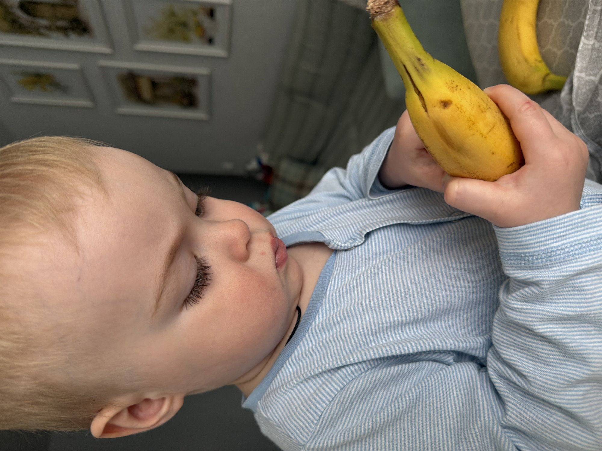 A close-up, slightly high-angle shot shows a baby holding a banana. The baby has fair skin, blonde hair, and closed eyes with long eyelashes. The baby is wearing a light blue and white striped shirt. The baby's hands are holding a ripe yellow banana. Another banana is partially visible in the upper right corner. In the blurred background, framed pictures are seen on a wall, and a grey curtain is present. The location is not specified, but the prompt states Votkinsk, Russia.