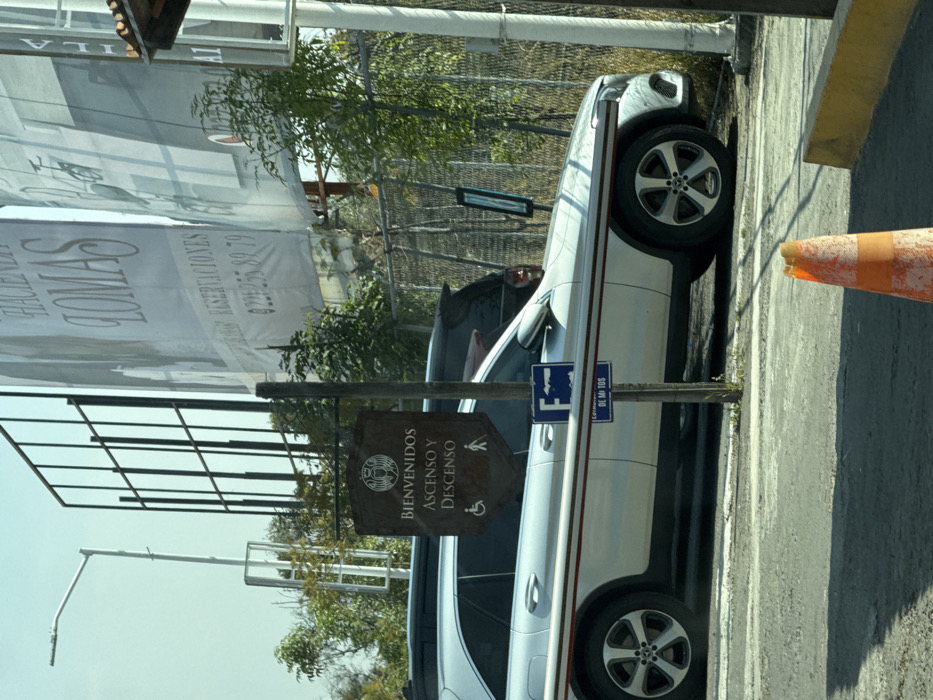 A white car parks adjacent to a pedestrian sign indicating "Ascenso y Descenso." The car, partially obscured, showcases its wheels and side profile. Behind, there's a large promotional banner alongside a metal fence. The ground is paved, featuring a traffic cone near the car. The sky appears clear, and trees are scattered in the background, providing a glimpse of greenery amidst the urban environment.