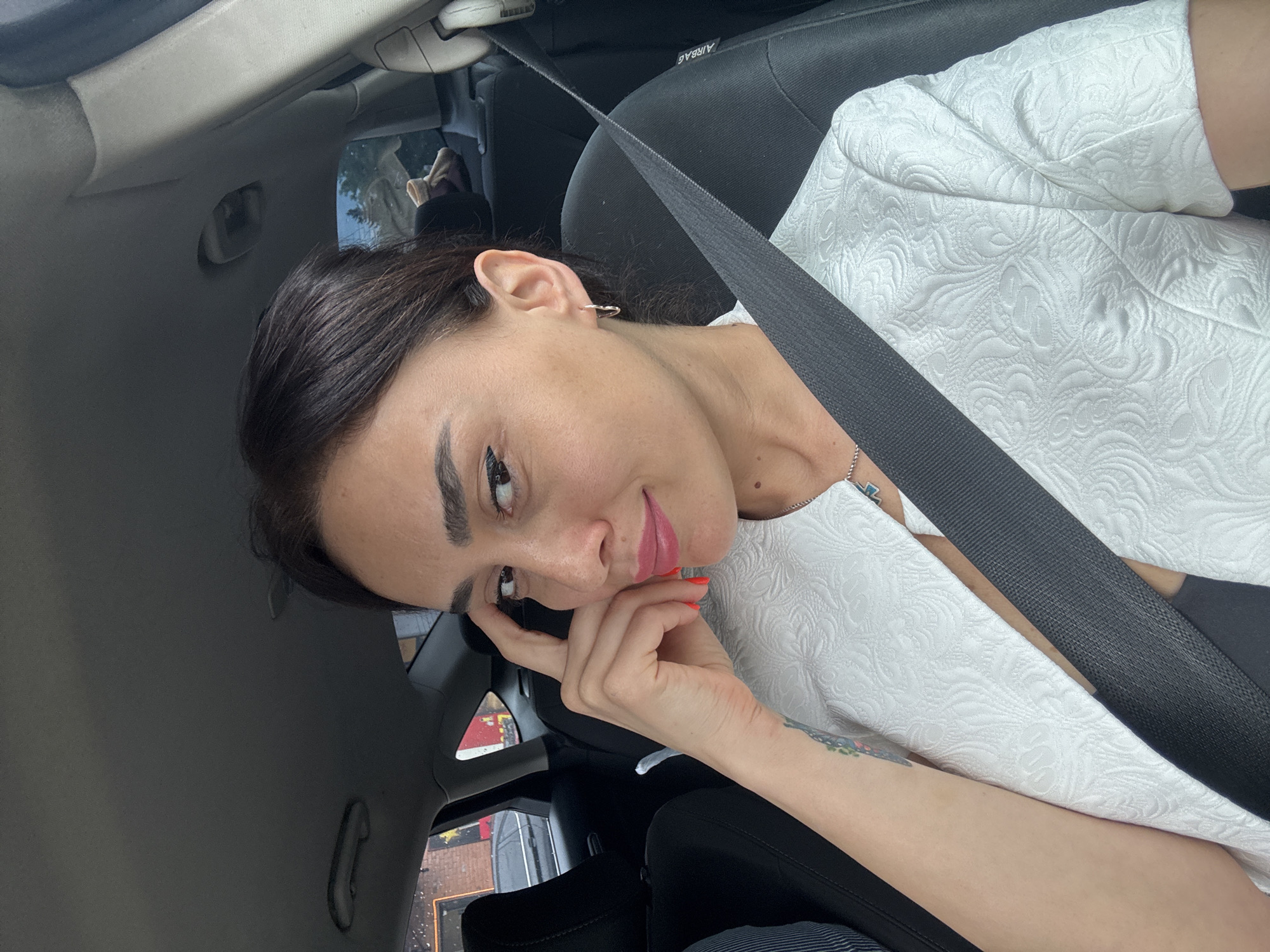 A woman with dark hair is in the passenger seat of a car, wearing a white textured jacket and a seatbelt. She is looking at the camera, with her right hand touching her forehead and left arm resting on her lap. The background shows the interior of a car, with seats and windows revealing a blurred view of the exterior, possibly a street scene in Tbilisi, Georgia.