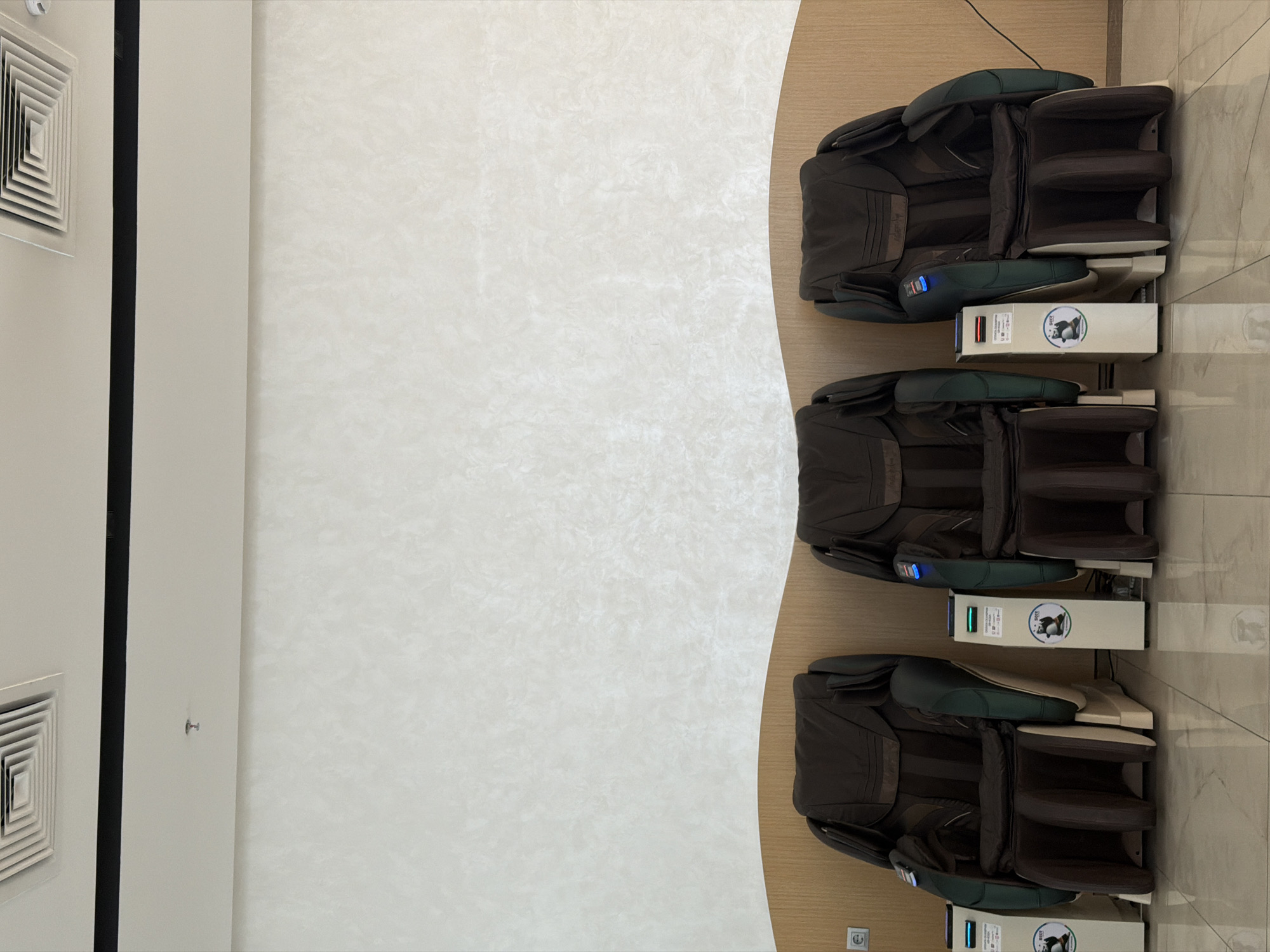 The image shows a row of three dark brown leather massage chairs with integrated control panels in front of them. The chairs are arranged in a well-lit interior space with a textured light-colored wall behind them. The floor is tiled with a reflective surface. A curved wooden panel separates the chairs from the wall. A black strip runs across the top of the wall, with a ventilation grate visible in the upper left corner. A small, silver fire sprinkler is visible on the ceiling. The control panels each have a digital display, a payment slot, and a circular sticker featuring a cartoon panda. The chairs are positioned to face forward, suggesting they are ready for use. Based on the presence of the panda sticker and the style of the massage chairs, this scene is likely located at a massage chair rental area within a public space in Tashkent, Uzbekistan, possibly an airport or a shopping mall.