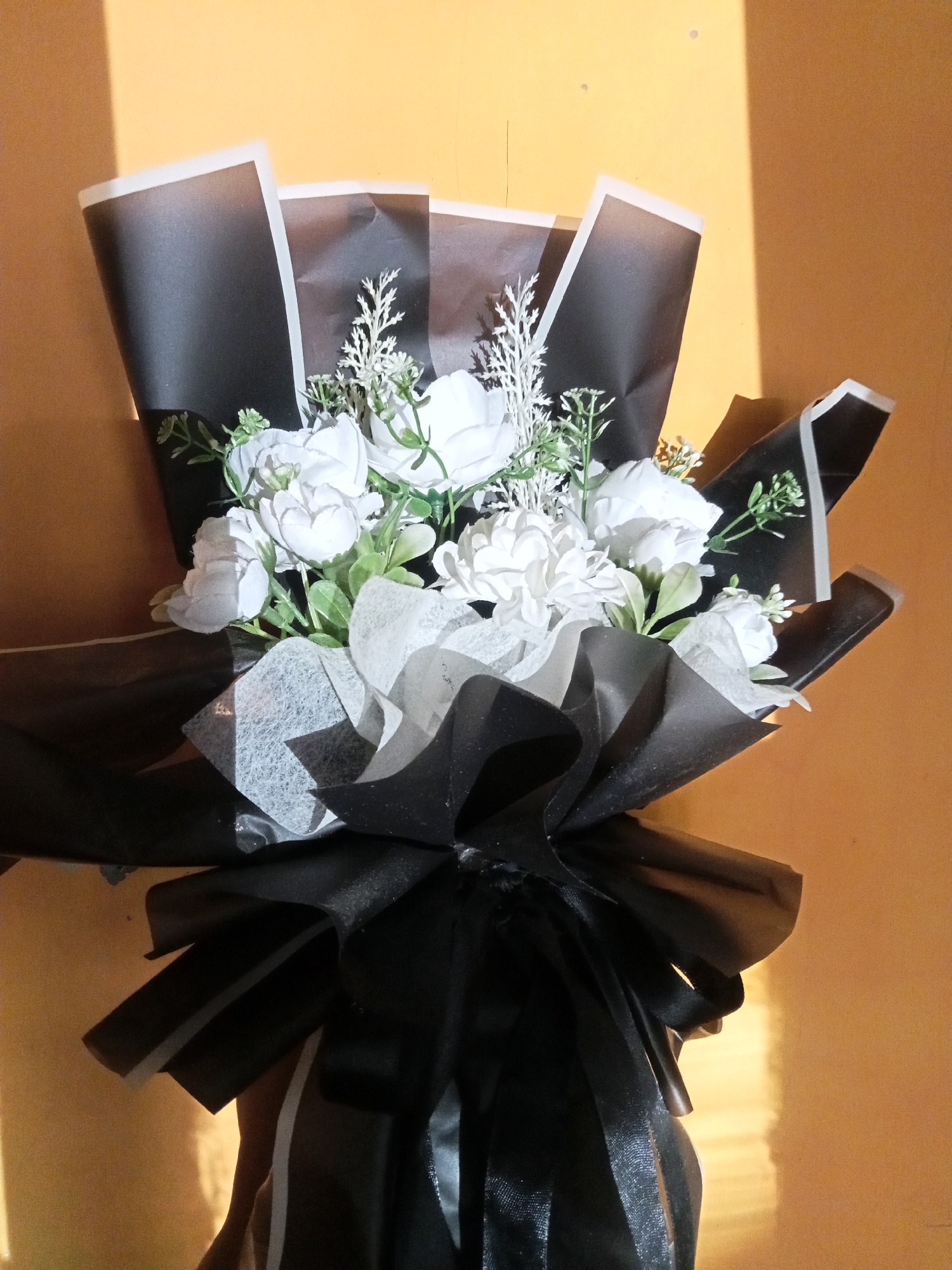 A bouquet of white artificial flowers with green leaves is presented against an orange wall, partially illuminated by sunlight casting a diagonal shadow. The bouquet is wrapped in black paper with white trim, and a black ribbon is tied in a bow at the base. The scene is likely captured indoors or in a sheltered outdoor area in Pancurwening, Indonesia, given the warm lighting and wall texture. There are no people or discernible activities depicted in the image.