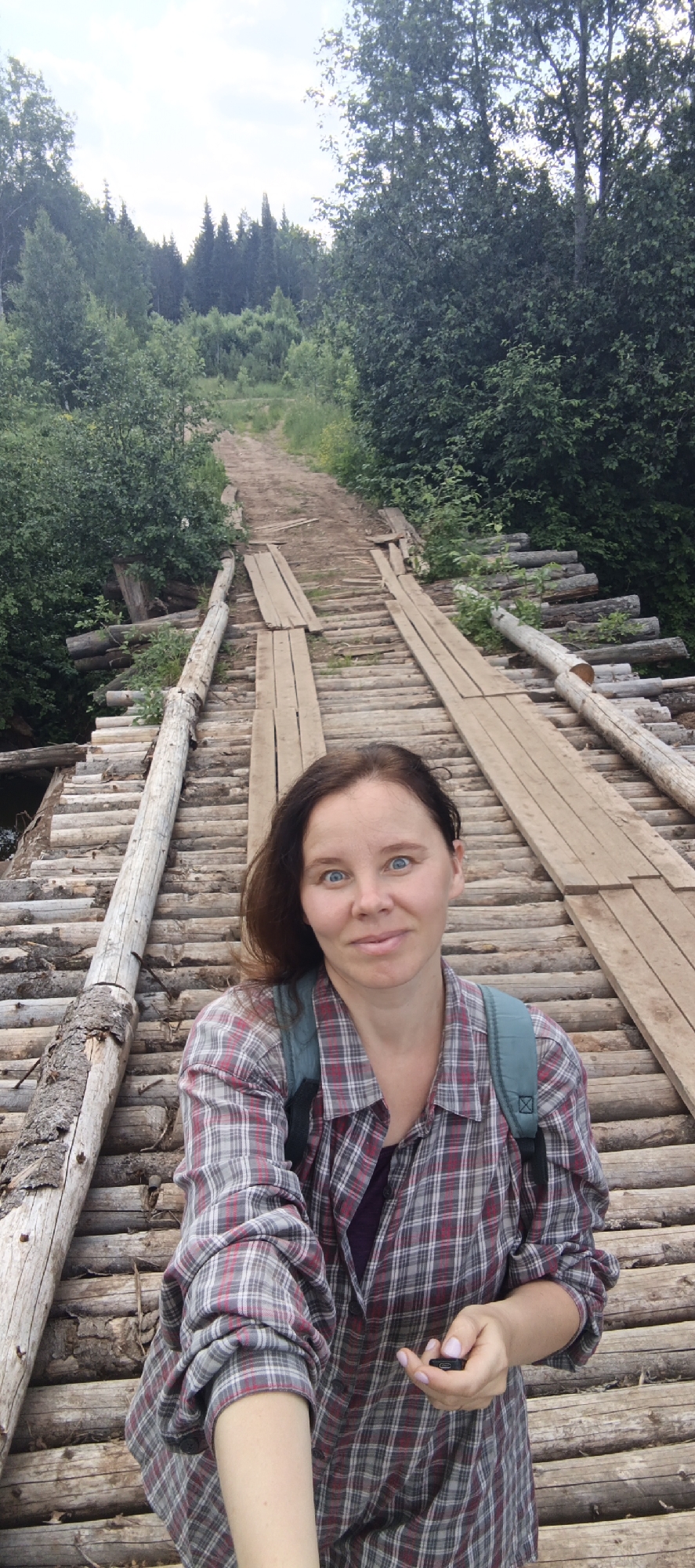 A woman with blue eyes and brown hair, wearing a red and gray plaid shirt and a teal backpack, takes a selfie on a wooden bridge. The bridge is constructed from logs and planks and spans across a wooded area. The background shows a dirt road leading uphill through a forest. The scene is outdoors, with lush green trees and vegetation.