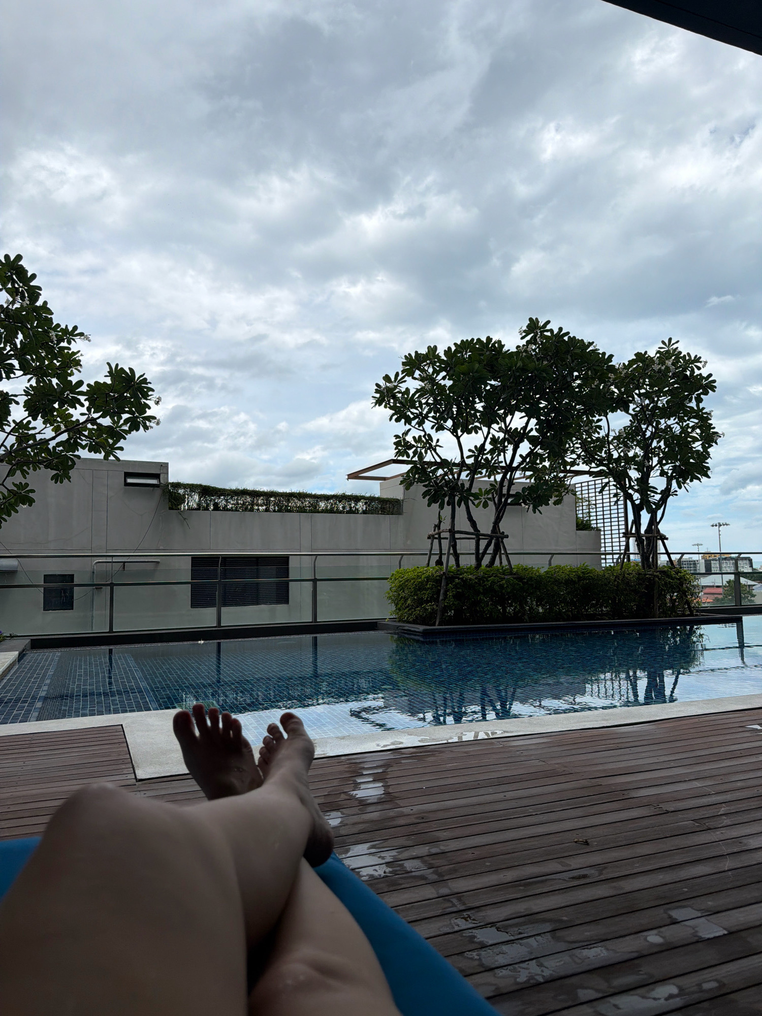The image depicts a person's legs and feet resting on a blue lounge chair on a wooden deck next to a swimming pool. The pool has tiled bottom and is reflecting the cloudy sky and surrounding trees. In the background, there is a modern building with a balcony and some greenery. The sky is overcast with gray clouds. The location is identified as Hua Hin City Municipality, Thailand. The overall scene suggests a relaxed atmosphere at a hotel or resort.