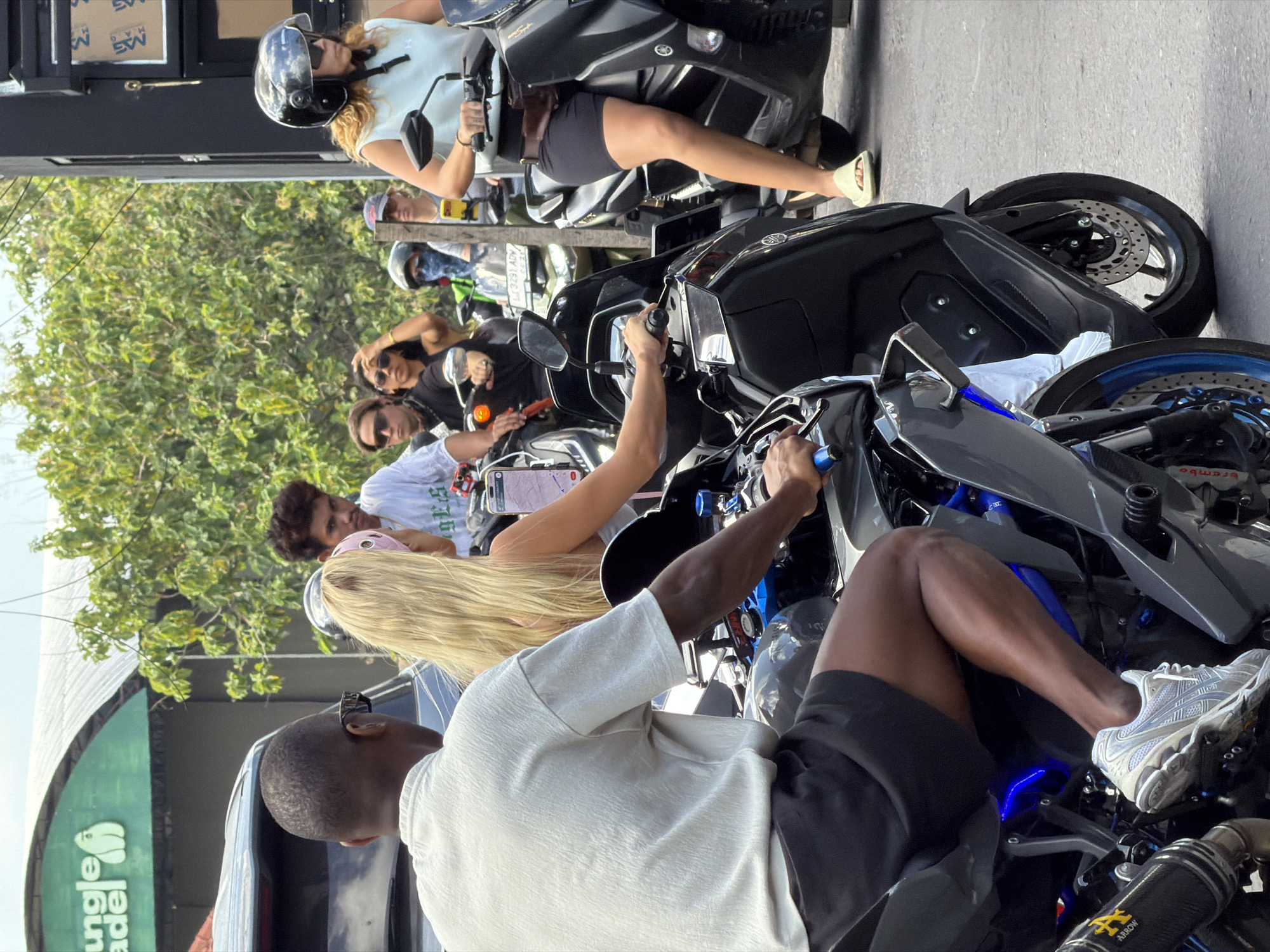 In North Kuta, Indonesia, on a bright day, a group of people on motorcycles are stopped in traffic. The foreground features a Black man wearing a light-colored shirt and dark shorts, with a blonde woman in a light blue tank top and dark shorts riding behind him. The woman is wearing a helmet and sunglasses and has a pink headscarf. Both are on a black motorcycle. To the right, another woman wearing a black helmet and sunglasses is riding a black motorcycle. She is wearing a light blue tank top and dark shorts, with tan slides on her feet. In the background, several other people on motorcycles are visible, including a person wearing a bandana over their face. Behind the motorcycles, lush green trees line the street. A portion of a building with a green awning and the text "Jungle" is visible on the left. The road surface is asphalt, and the lighting suggests it is daytime with sunlight.