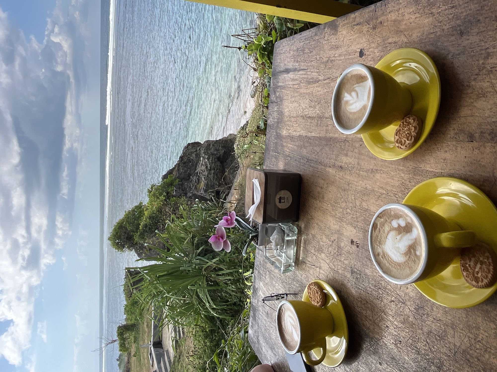 A high-angle, eye-level shot shows two yellow cups of latte with foam art and cookies on yellow saucers placed on a wooden table. The table also holds a pair of glasses, an ashtray with sugar packets, a small potted orchid with pink flowers, and a wooden tissue box. The table is positioned on a balcony overlooking a cliff with lush green vegetation and the ocean. White waves crash on the shore below, and the sky is filled with scattered clouds. This scene is located at a cliffside cafe in Kuta Selatan, Indonesia, known for its stunning ocean views.