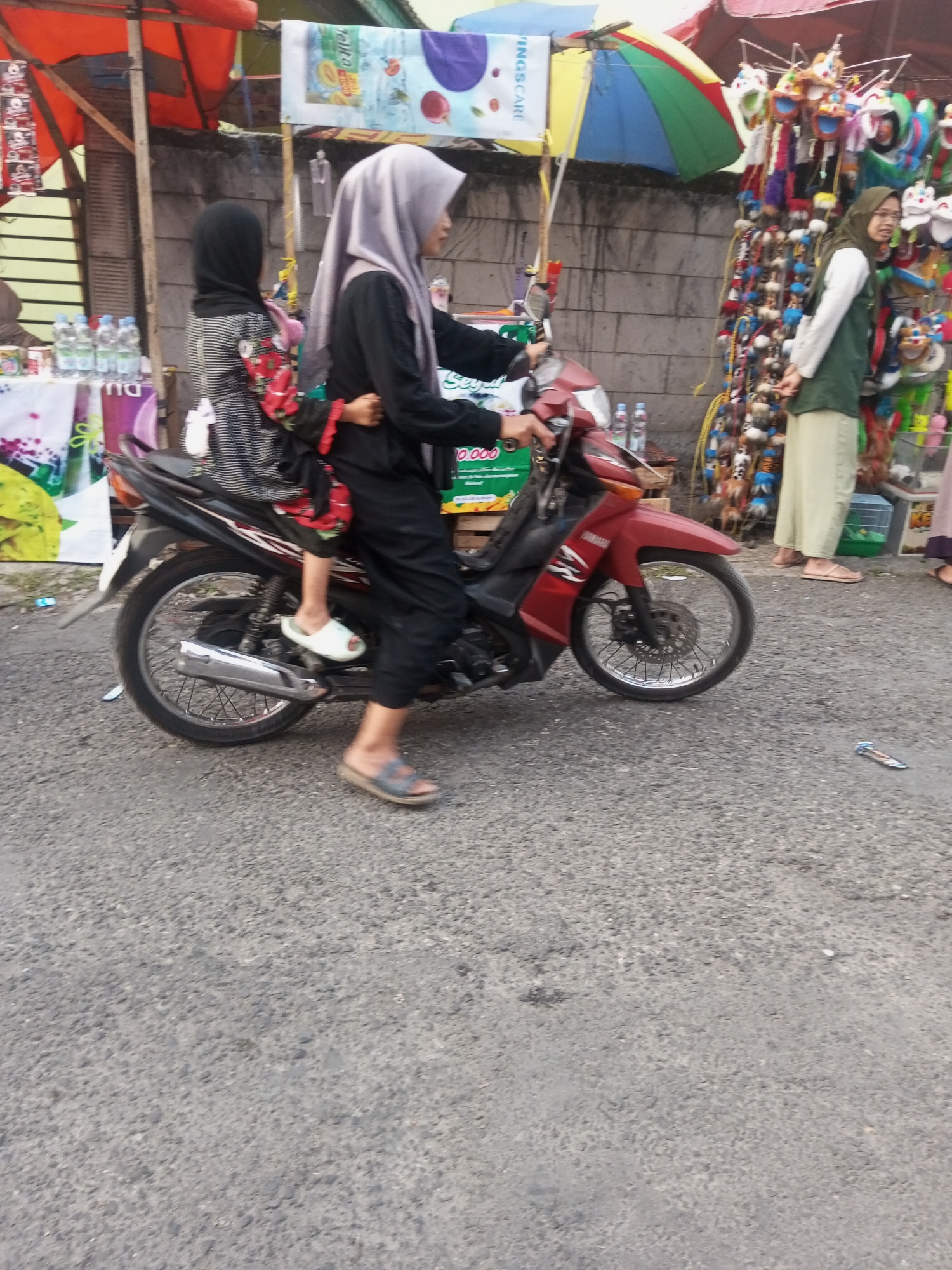 In Pandansari, Indonesia, two individuals are depicted on a motorcycle on a paved road. The rider, wearing a gray hijab and black clothing, is controlling the motorcycle. Behind the rider, a passenger wearing a black hijab and a patterned dress is seated. The motorcycle is red and black. In the background, a market scene is visible with stalls displaying various goods, including umbrellas, toys, and bottles of water. The overall environment appears to be an outdoor market.