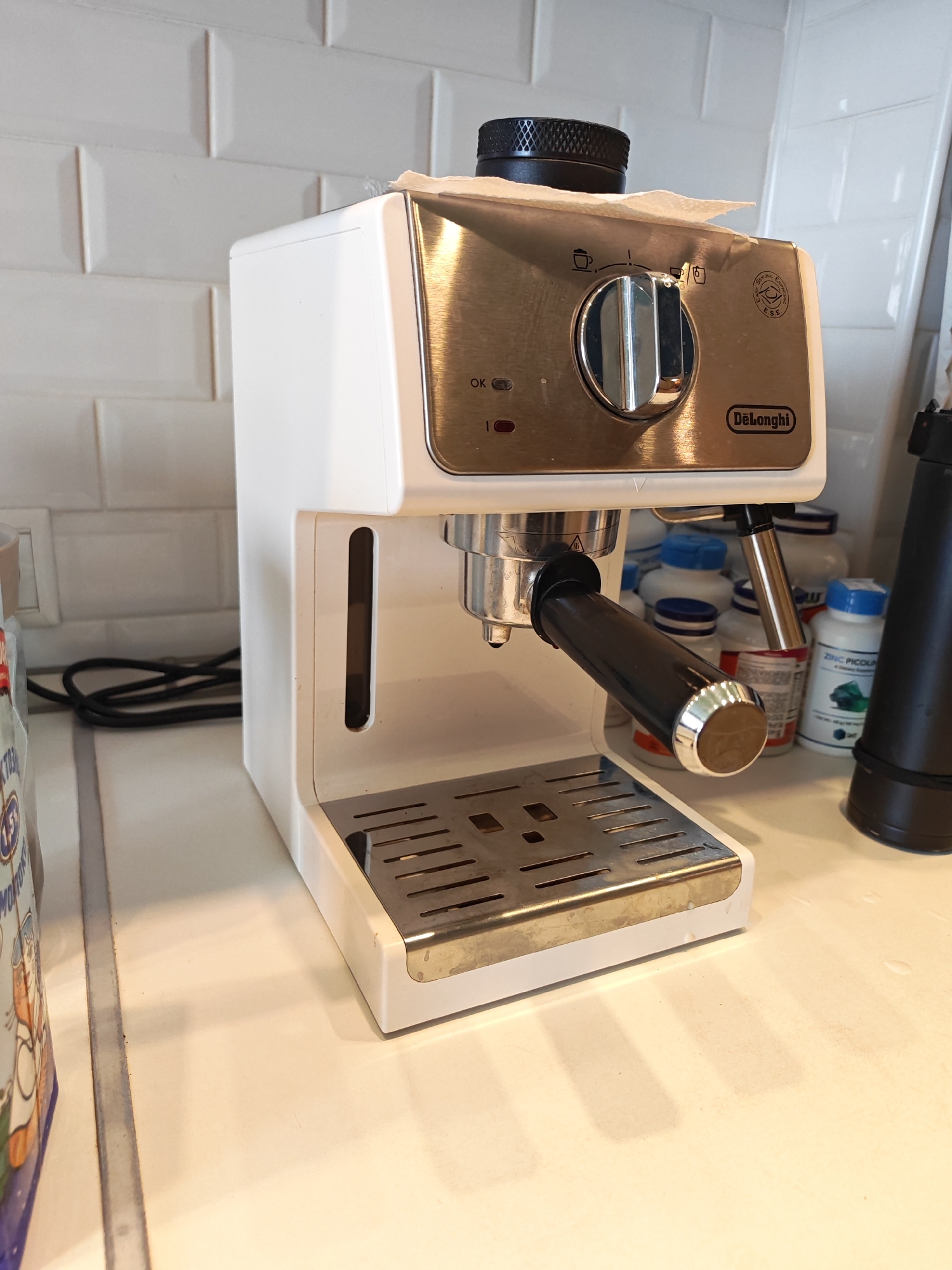 An espresso machine sits on a white countertop, in front of a white tiled backsplash. The espresso machine is white and stainless steel, with a black portafilter. To the right of the espresso machine is a black object, possibly a coffee grinder or a frother. In the background, there are several bottles of supplements. To the left of the espresso machine is a portion of a package with a cartoon image.