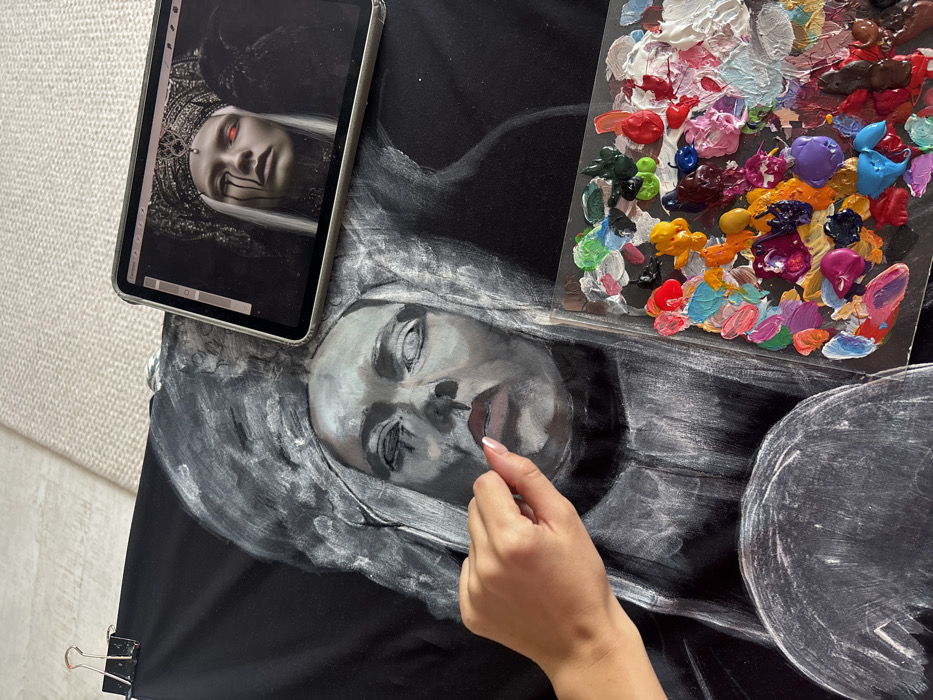 The scene depicts an art creation process occurring in the city of Krasnoyarsk, Russia. The primary subject is an overhead view of a large black canvas, upon which a grayscale portrait of a woman with an elaborate headdress is being painted. Her face features shadowed eyes and partially rendered red lips.
A human left hand, with manicured fingernails, is positioned over the canvas near the portrait's mouth, appearing to apply or blend paint. A clear, rectangular artist's palette, heavily covered with numerous distinct blobs of paint in various colors including red, pink, orange, yellow, green, blue, purple, white, black, and brown, rests in the lower right portion of the frame, overlapping the canvas.
In the upper right, a digital tablet, likely silver in color, displays a reference image. This image shows a woman with pale skin, white hair, a dark, ornate crown, red eyes, and dark liquid streaks below her eyes. A dark, avian-like figure is visible to the right of the reference image woman.
The canvas is secured at the top left by a black binder clip with a red handle. The background beyond the immediate art materials consists of a light-colored, textured surface, possibly carpet or a rug, indicating an indoor setting.