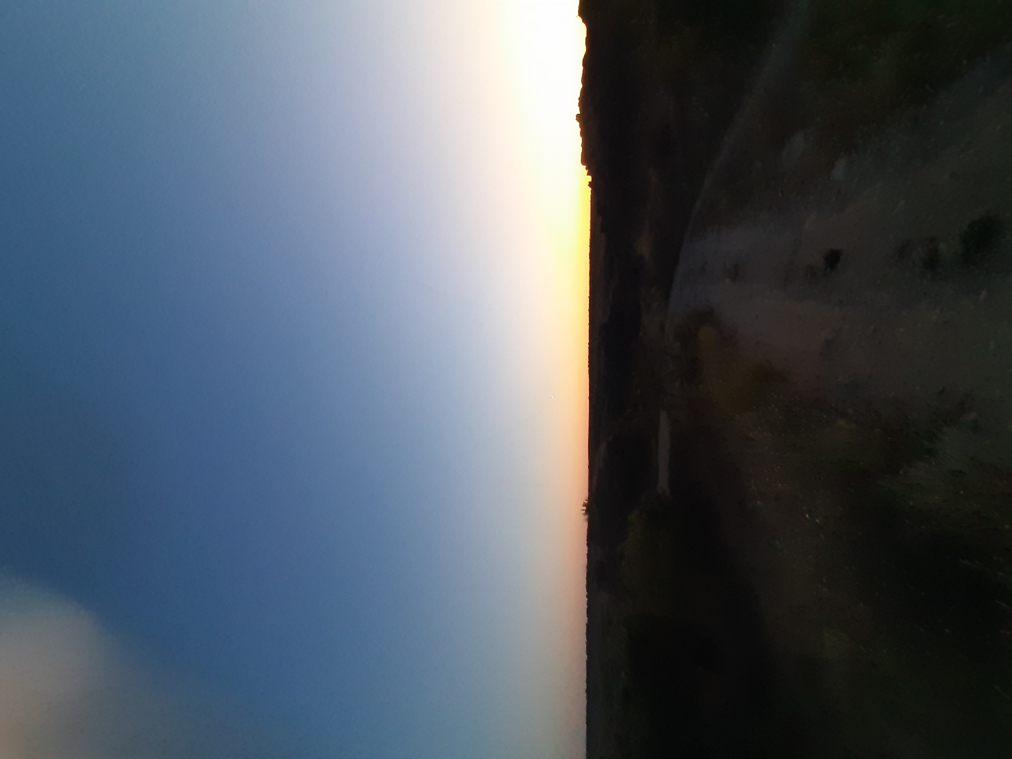 The image depicts a rural landscape at dusk. A dirt road curves through dark, low-lying vegetation in the foreground, leading towards the horizon. The sky transitions from a deep blue at the top to a pale orange near the horizon, indicating the setting or rising sun. Silhouetted against the horizon are low hills or rock formations, with a hint of light from beyond. The overall impression is one of solitude and the transition between day and night in a natural environment. The location is identified as Unknown, Unknown.