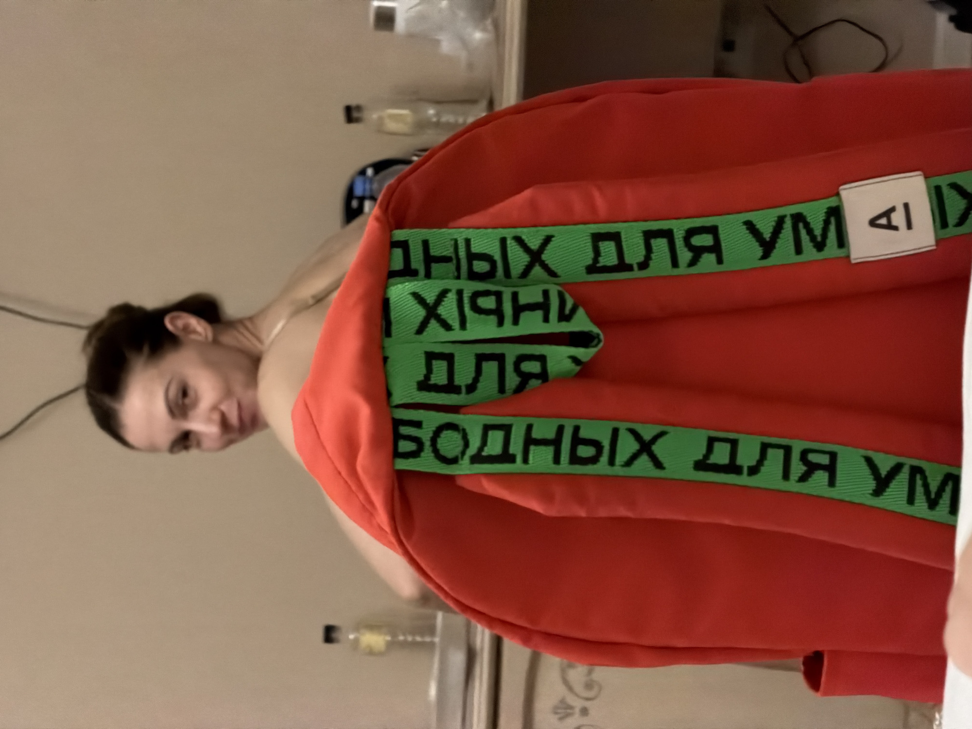 An adult female with her hair in a bun is sitting facing away from the camera, looking over her left shoulder with a slight smile. She is wearing a strapless garment. She is partially obscured by a bright red backpack, which has two green straps with black Cyrillic lettering. A small white tag with the letter "A" is attached to the right strap. The background appears to be an indoor setting, possibly a bedroom, with a wall, a dresser, and a shelf visible.
