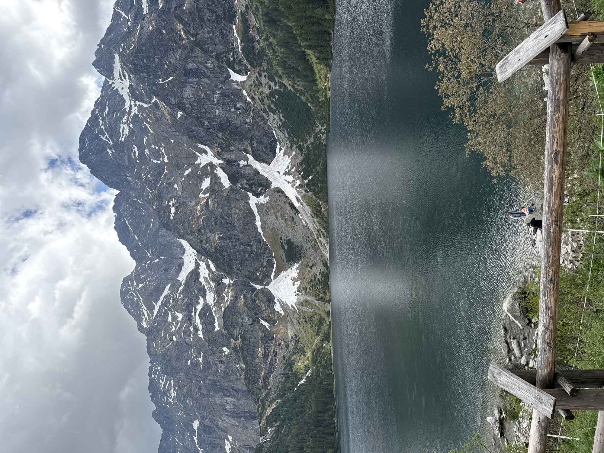 The image captures a mountain lake scene. A large, craggy mountain with snow patches dominates the left side of the frame, under a cloudy sky. The right side features a dark, still lake reflecting light. A wooden railing is visible in the foreground, partially obscuring the shoreline. People are on the edge of the lake. This scene could be in the city of Brzegi, Poland.