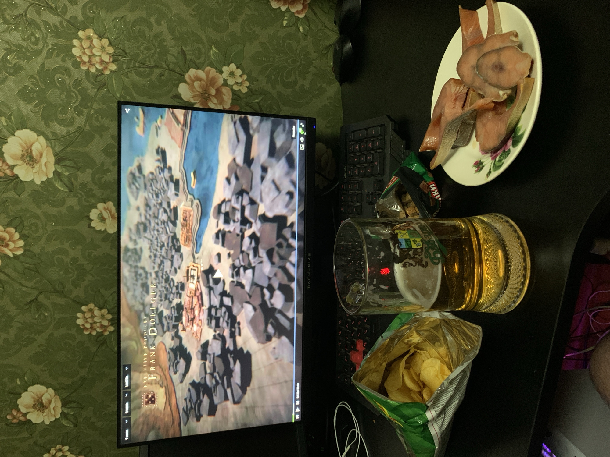 An overhead shot shows a computer monitor displaying a video game map. The game map is of a city with various buildings and a body of water. The computer monitor is placed on a black desk, with a keyboard positioned in front of it. To the left of the monitor, a bag of potato chips is partially opened, with chips spilling out. To the right of the chips, a glass mug filled with beer sits on the desk. Behind the mug, on a white plate adorned with a floral pattern, are several slices of cooked fish. The background of the image is a wall covered in green floral wallpaper. There are no people visible in the image, and the scene appears to be a casual setting, possibly a gaming session or a meal. The location of Desyatikhatka, Russia is not depicted or indicated in the image.