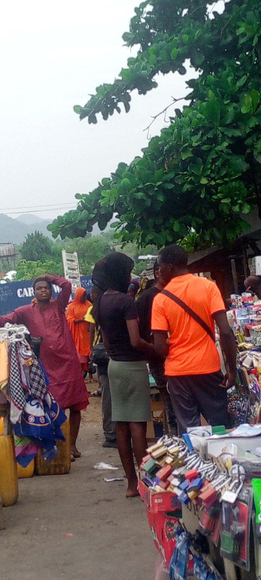 The image depicts a street scene in Adankolo, Nigeria. Several people are visible, including a man in a maroon garment standing near a display of scarves and bags. Another person is wearing an orange shirt and carrying a bag. A woman in a black shirt and green skirt stands next to him. The scene includes street vendors selling various goods such as locks, accessories, and household items. A large tree is visible in the background, and there are some hills or mountains in the distance, under an overcast sky. A partially visible sign says "CAR WASH". The ground appears to be paved or made of compacted earth.