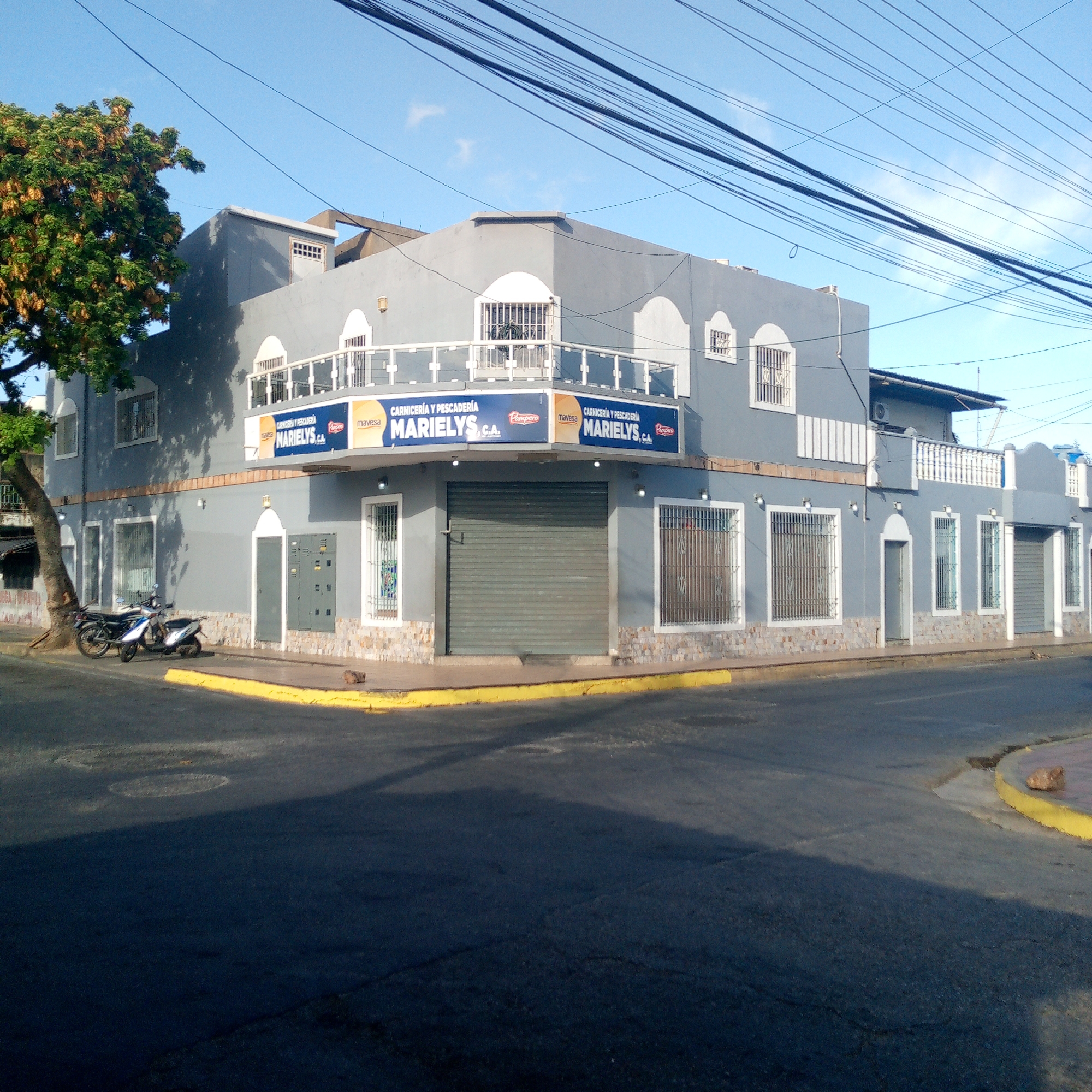 The image shows a two-story gray building, likely a commercial establishment, with signage indicating "CARNICERÍA Y PESCADERÍA MARIELYS, C.A.". The building has a facade with arched windows, some protected by metal bars. A prominent blue sign with white text and logos is visible above the ground floor windows, which suggests a meat and fish market. A large roll-up metal door is closed on one corner of the building. Two motorcycles are parked next to the building, near a tree. Overhead, numerous power lines crisscross the sky. The building is located at an intersection with yellow-painted curb. The scene appears to be in Juan Griego, Venezuela.