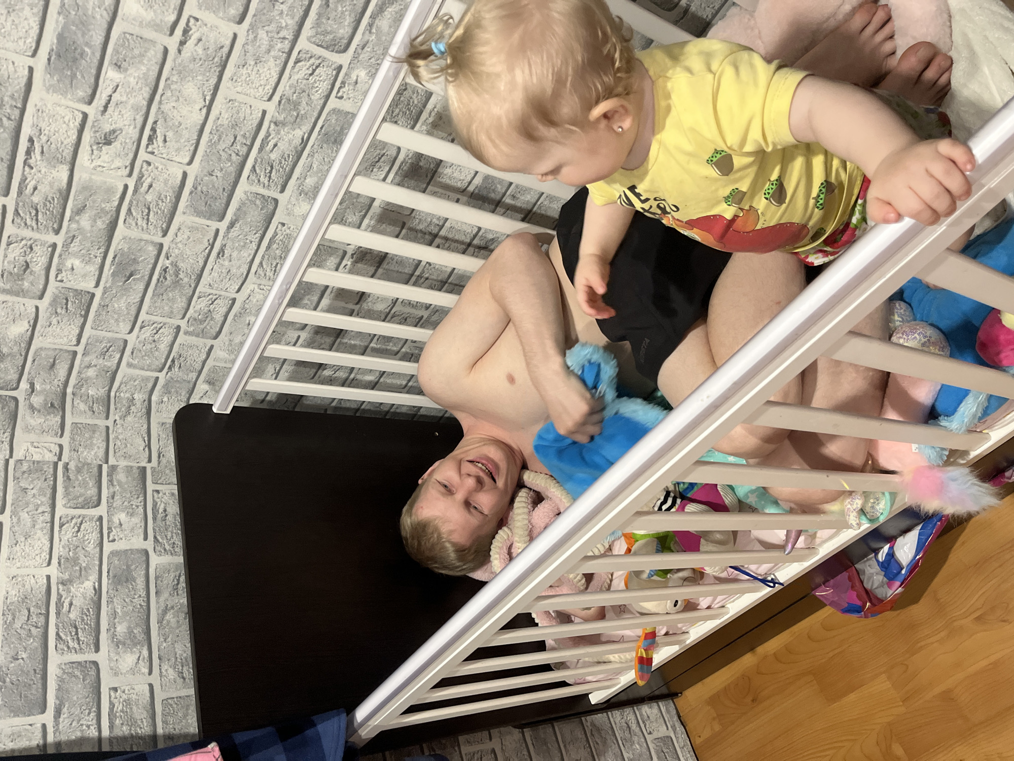 A fair-skinned adult male with blond hair is lying on his back in a white crib, holding a blue plush toy. A young, fair-skinned child with blond hair stands at the edge of the crib, wearing a yellow t-shirt with cartoon images and dark-colored shorts. The room has a faux gray brick wall and wood flooring. The crib contains multiple plush toys and bedding. The image's origin or location in Vesyoloye, Russia, is not evident from visual information.