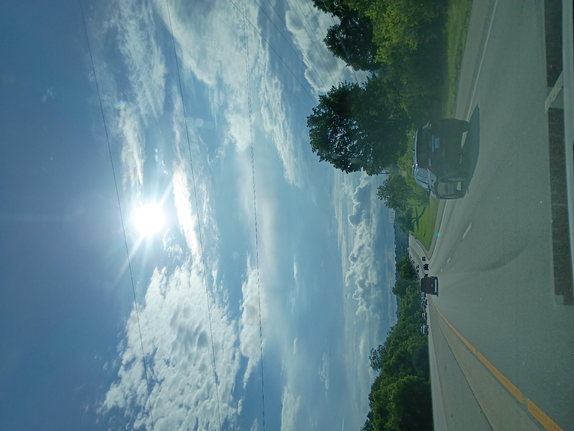 An eye-level, outdoor, medium shot captures a highway scene under a bright, sunlit sky with scattered clouds. The sun, positioned high and slightly to the left, illuminates the sky, casting bright light and some lens flare. The highway stretches into the distance, with vehicles moving in both directions. In the foreground, a black SUV is visible on the right lane, heading away from the viewer. The road is flanked by lush green trees on both sides, with a few power lines crossing the sky horizontally. The overall scene conveys a sense of travel and a sunny day on the road. The location is the city of Lexington, United States of America.