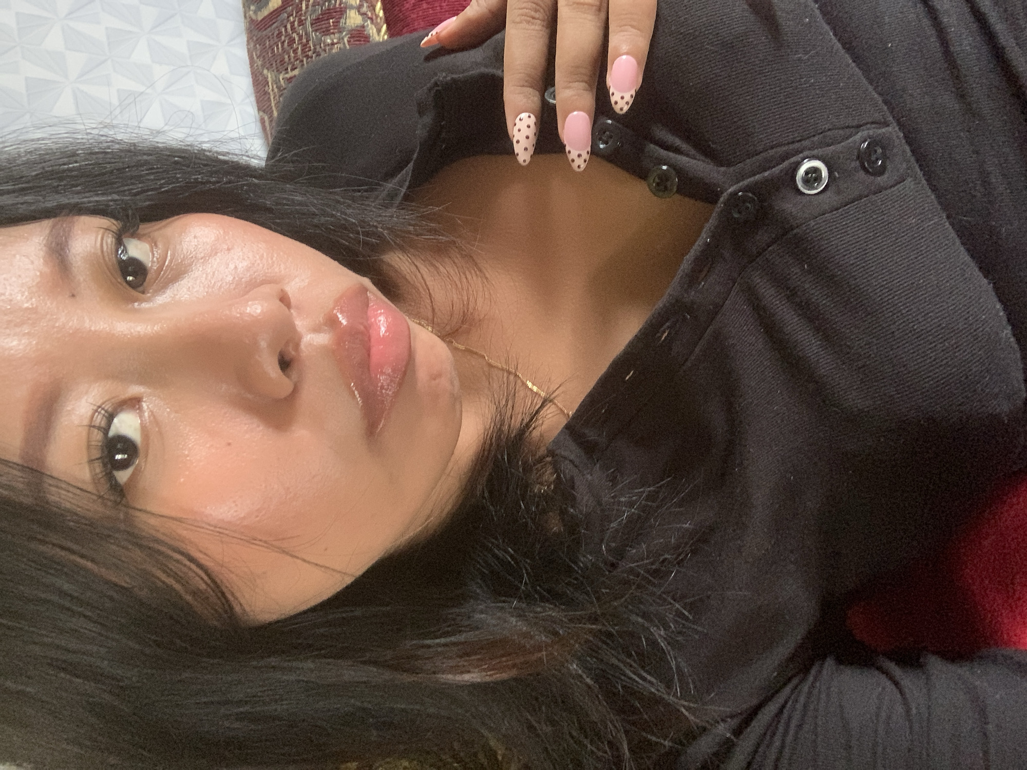 A close-up, high-angle, selfie-style, medium shot shows a person with dark hair, fair skin, and full lips, wearing a black, collared shirt with buttons down the placket. The person has long, dark hair that falls loosely around their face and shoulders. Their eyes are dark and looking towards the camera. Their lips are glossy and appear to be slightly parted. A thin gold chain necklace is visible around their neck. The person's right hand, with manicured nails featuring a pink base with black polka dots, is resting on their chest, near the buttons of the shirt. The background is out of focus, showing a patterned wallpaper and a hint of a textured fabric in red and gold. There is no discernible information about the location in Jajag, Indonesia.