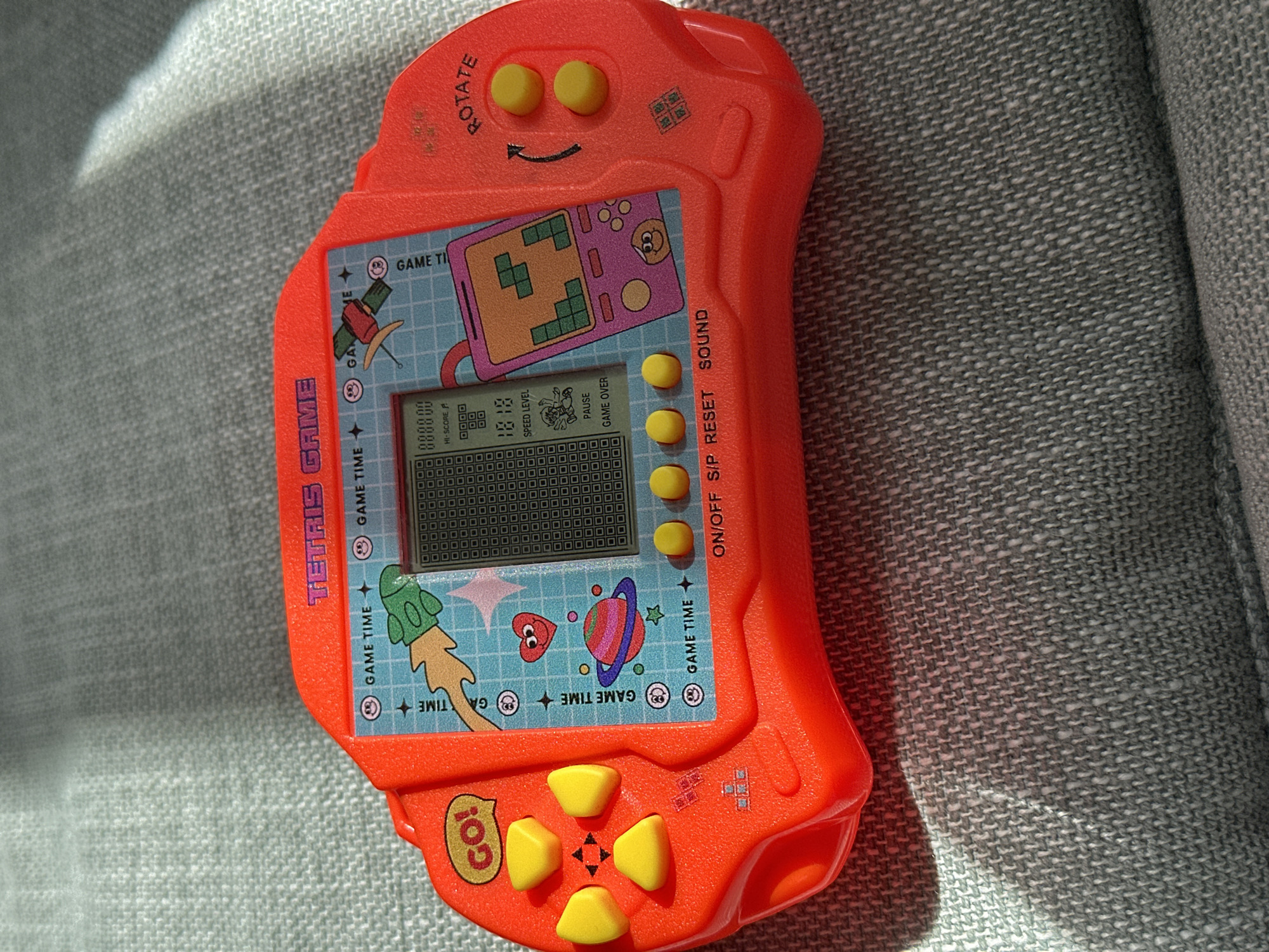 A red, handheld electronic game, likely a Tetris clone, rests on a grey, woven fabric surface. The game console features a screen displaying a Tetris game in progress, along with game time, score, speed level, pause, and game over indicators. It has a directional pad on the left with a "GO!" button and two yellow action buttons on the right labeled "ROTATE." The console is adorned with cartoonish graphics including rockets, planets, and smiley faces. The fabric surface appears to be upholstery, possibly from a sofa or chair. The lighting suggests it is daytime, with sunlight casting shadows. There is no indication of the specific location in Moortown, Ireland; the scene is isolated to the object and its immediate surroundings.