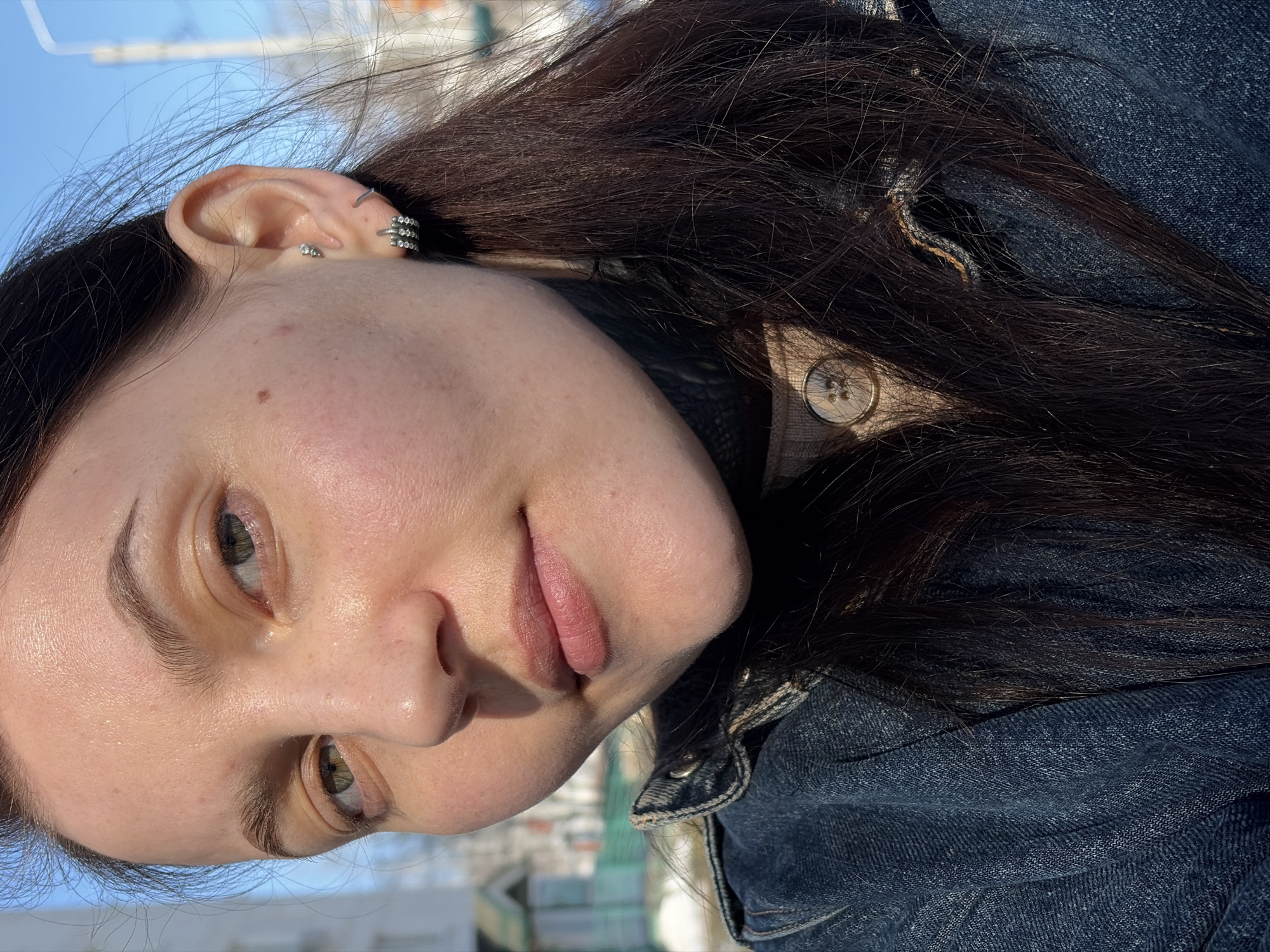 A close-up view depicts a female individual outdoors in Chaikovsky, Russia. Her face, angled slightly, is illuminated by direct sunlight originating from the left, highlighting her fair skin, light green eyes, and light pink, closed lips. She has dark brown, long hair cascading over her shoulders and chest. Her visible right ear exhibits multiple piercings: a small stud in the upper helix, a thin silver cuff in the mid-helix, and a larger, multi-row silver-toned hoop earring in the lobe. She is dressed in a dark blue denim jacket worn over a beige knit garment with a visible round button. The background, under a clear blue sky, reveals parts of a light-colored building to the left, a street light pole to the right, and a more distant house with a peaked roof and a green fence. Patches of snow are visible on the ground, indicating a cold, sunny day.
