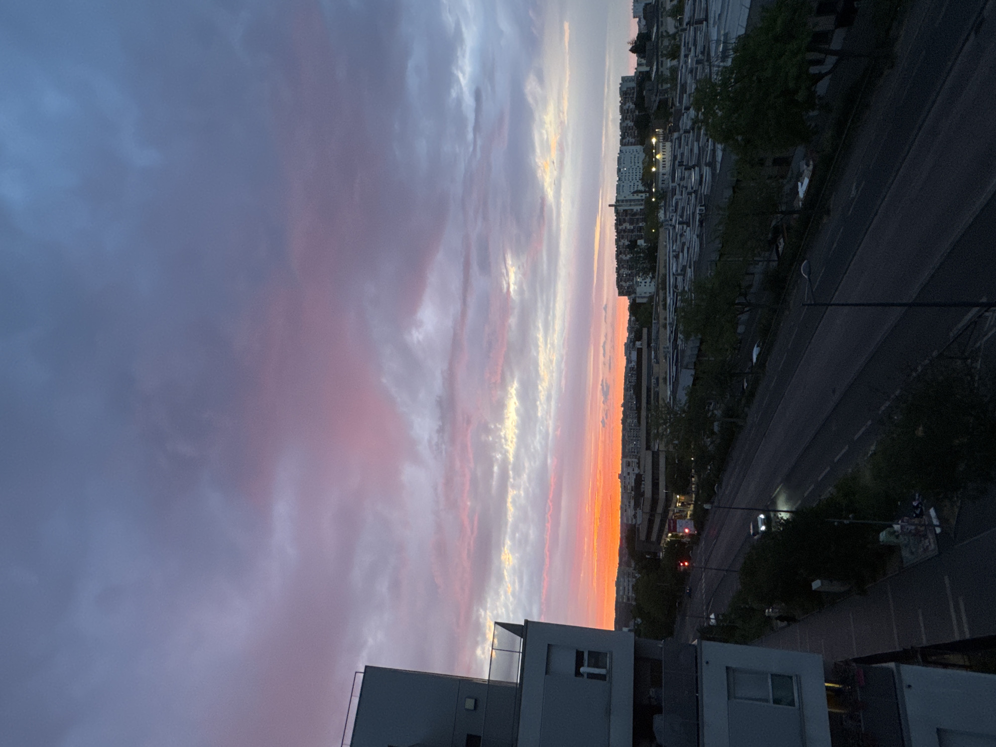 The image depicts a city skyline at dusk, characterized by a dramatic sky painted with hues of purple, pink, and orange. The foreground features the corner of a modern apartment building with a glass-paneled balcony. Below, a road runs through the urban landscape, with a few cars visible, one with its headlights on. The middle ground is dominated by a collection of low-rise buildings and industrial structures with flat roofs, interspersed with trees. In the distance, a cluster of taller apartment buildings forms the backdrop, with some windows illuminated. The overall scene is one of an urban environment transitioning from day to night, with the vibrant sky serving as a striking visual element.