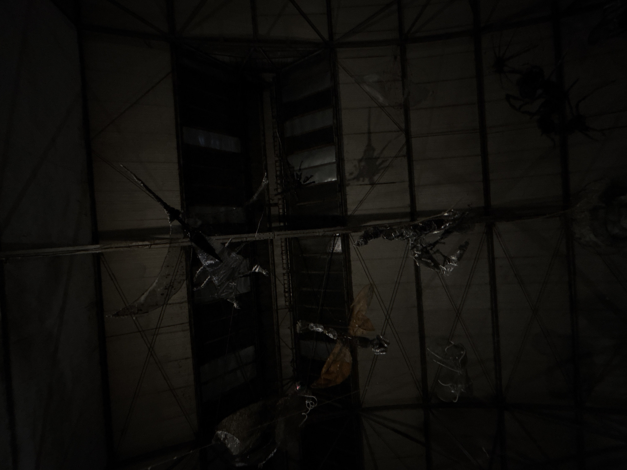 The image depicts the interior of an industrial or abandoned building in Belgrade, Serbia. The scene is dark, illuminated only by faint light filtering through openings. The ceiling features a grid of metal beams and panels. Suspended from the ceiling are various indeterminate objects, possibly debris, decorations, or installations. Their shapes are unclear due to the low light. The overall impression is one of neglect and disuse. No people are visible.