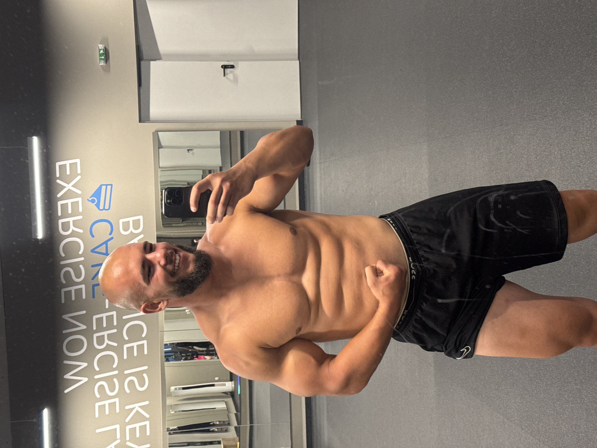 The image shows a man with a bald head and beard posing for a mirror selfie in a gym. He is wearing black athletic shorts. The gym appears to be modern, with grey walls and floors, and bright fluorescent lighting. In the background, there are mirrors, gym equipment, and white doors. A motivational phrase is visible on the wall behind the man, reading "EXERCISE IS KEY TO CARE-FREE LIFE." The setting is the Aura Fitness gym located in Sofia, Bulgaria. The man is flexing his bicep and showing his physique.