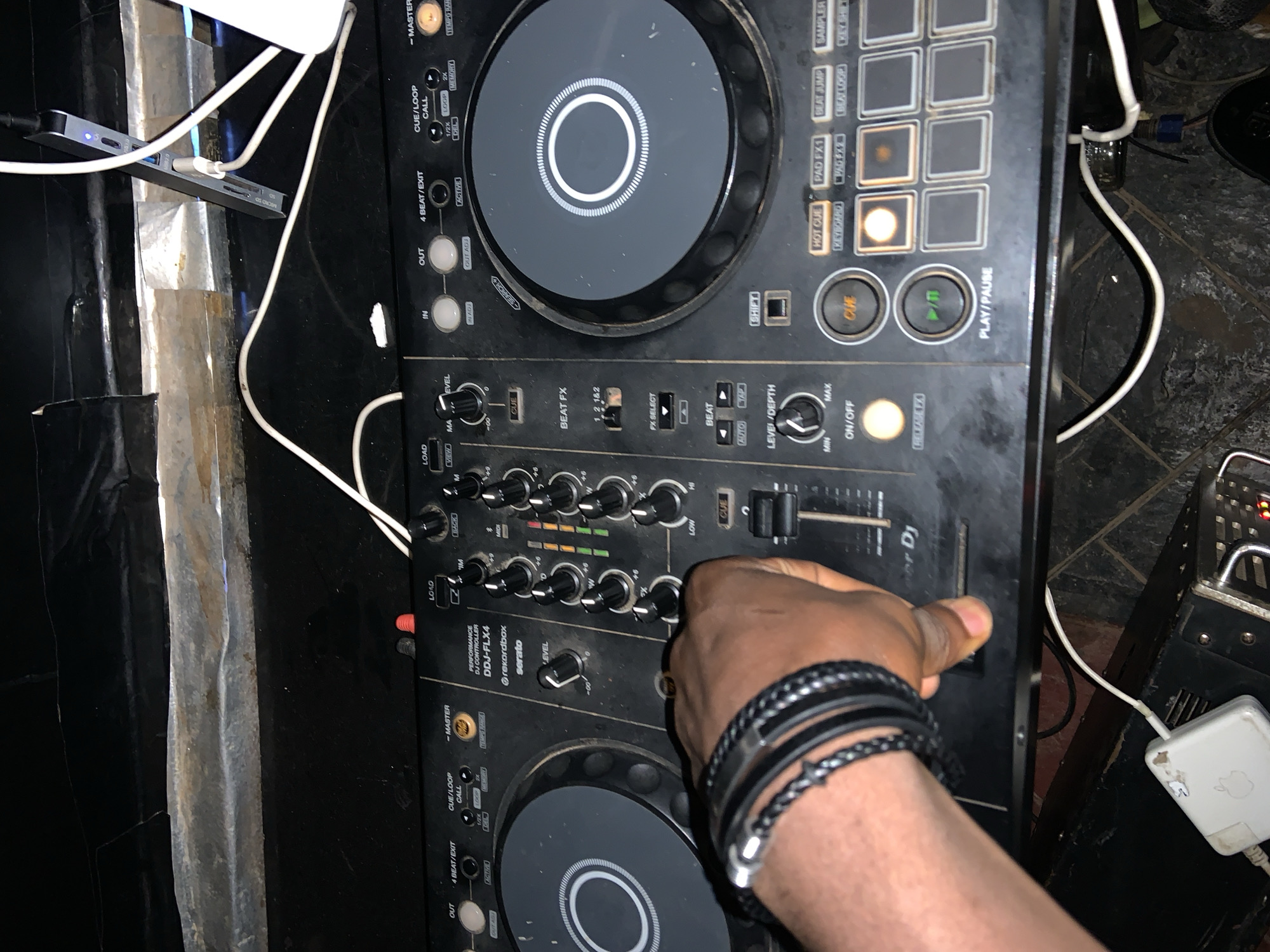 The image shows a close-up of a Pioneer DJ controller, model DDJ-FLX4, being operated by a person's hand. The controller is black and has various knobs, buttons, and illuminated pads. A hand wearing black braided bracelets is visible, with its fingers manipulating a slider on the left side of the controller. The controller is placed on a black surface, possibly a table or stand, and is connected to power and other equipment via cables. In the background, there is a rough, metallic-looking surface with some black covering. The overall environment appears to be dimly lit, suggesting a performance or practice space. The setting is identified as Gura Top, Nigeria.