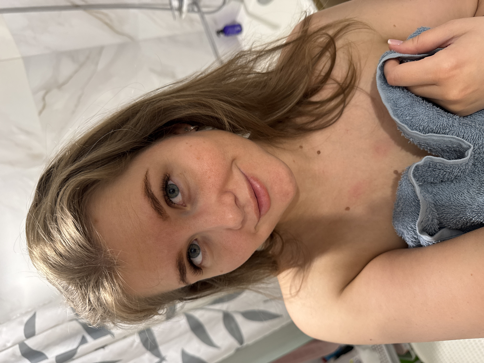 A woman with light brown hair and blue eyes is holding a light blue towel. She is in a bathroom, which is likely in Ферма, Russia. The bathroom has a white and grey marble-like wall and shower curtain. A shower head and various bottles are visible in the background.