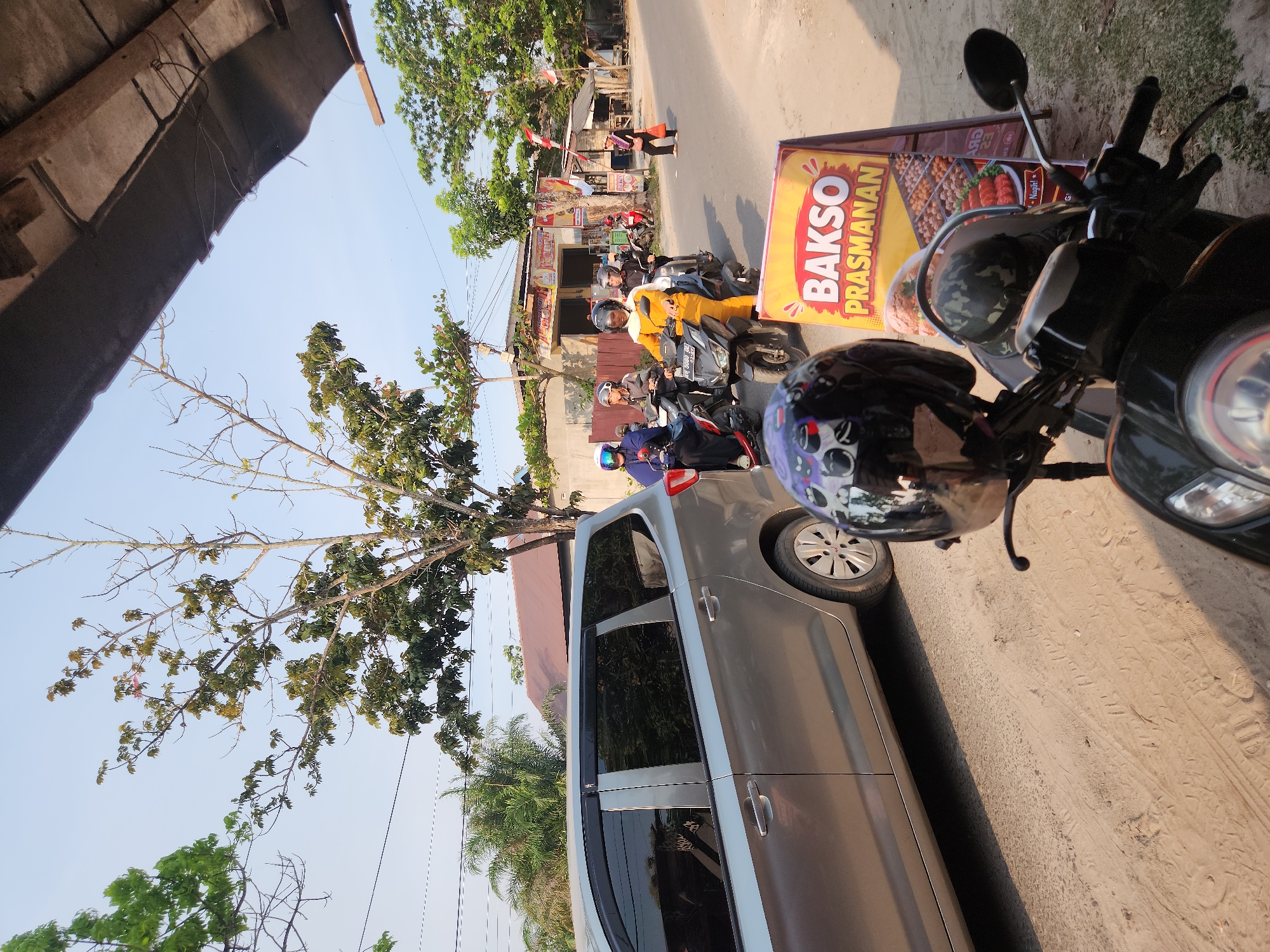 This is a ground-level, eye-level, full-color photograph captured on a sunny day in Sampit, Indonesia. The scene depicts a street with several motorcycles carrying riders and passengers, a silver minivan, and a prominent signboard advertising "Bakso Prasmanan".
In the foreground, a black motorcycle is parked, with a helmet placed on its handlebars. A second, identical helmet is visible on the back of the motorcycle. To the right of the motorcycle, a bright yellow A-frame signboard displays "BAKSO PRASMANAN" in large red letters with a shadow effect. Below the text, a grid of images showcases various food items, likely related to bakso (meatball soup).
Behind the motorcycle and signboard, a silver minivan is parked parallel to the street. Its windows are tinted, and the vehicle appears to be stationary.
Several motorcycles are in motion on the street, heading away from the viewer. The lead motorcycle has a rider wearing a white helmet and a dark blue garment. Behind them, another motorcycle has two occupants; the rider is wearing a grey helmet and a dark top, and the passenger wears a darker helmet and a light-colored jacket. The third motorcycle in the line has a rider in a bright yellow outfit and a black helmet.
Further down the street, more buildings and shops are visible. A woman in a purple headscarf and black clothing is walking away from the camera. A red and white Indonesian flag is mounted on a pole near one of the buildings. The environment is dusty and unpaved, with a clear blue sky overhead. Trees with sparse foliage are scattered throughout the scene. The overall impression is of a typical street scene in a Southeast Asian city.