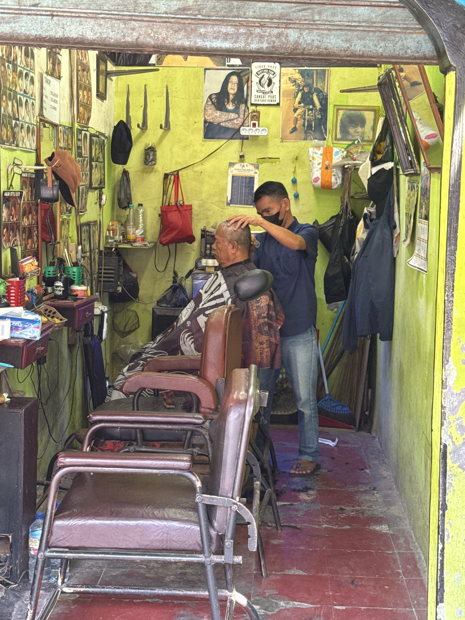 In a cramped, brightly lit barbershop in Sukaraja, Indonesia, a barber with short dark hair and a black face mask is trimming the hair of an elderly man. The barber, wearing a navy blue shirt and light blue jeans, uses his right hand to hold the man's head steady while his left hand is obscured by the man's hair. The elderly man, seated in a brown leather barber chair, is wearing a dark barber cape patterned with white and black designs.
The shop's walls are painted a vivid lime green and are adorned with numerous framed pictures of hairstyles, some faded. A collection of hair products and tools are visible on shelves and a small wooden table. A red fabric bag hangs on a hook on the wall, and a broom leans against the wall near a collection of clothing items. The floor is tiled in a reddish-brown hue, with scattered hair clippings visible. Two other barber chairs, one in the foreground and one behind the barber's chair, are empty. The overall impression is of a busy, functional, and well-worn neighborhood barbershop.