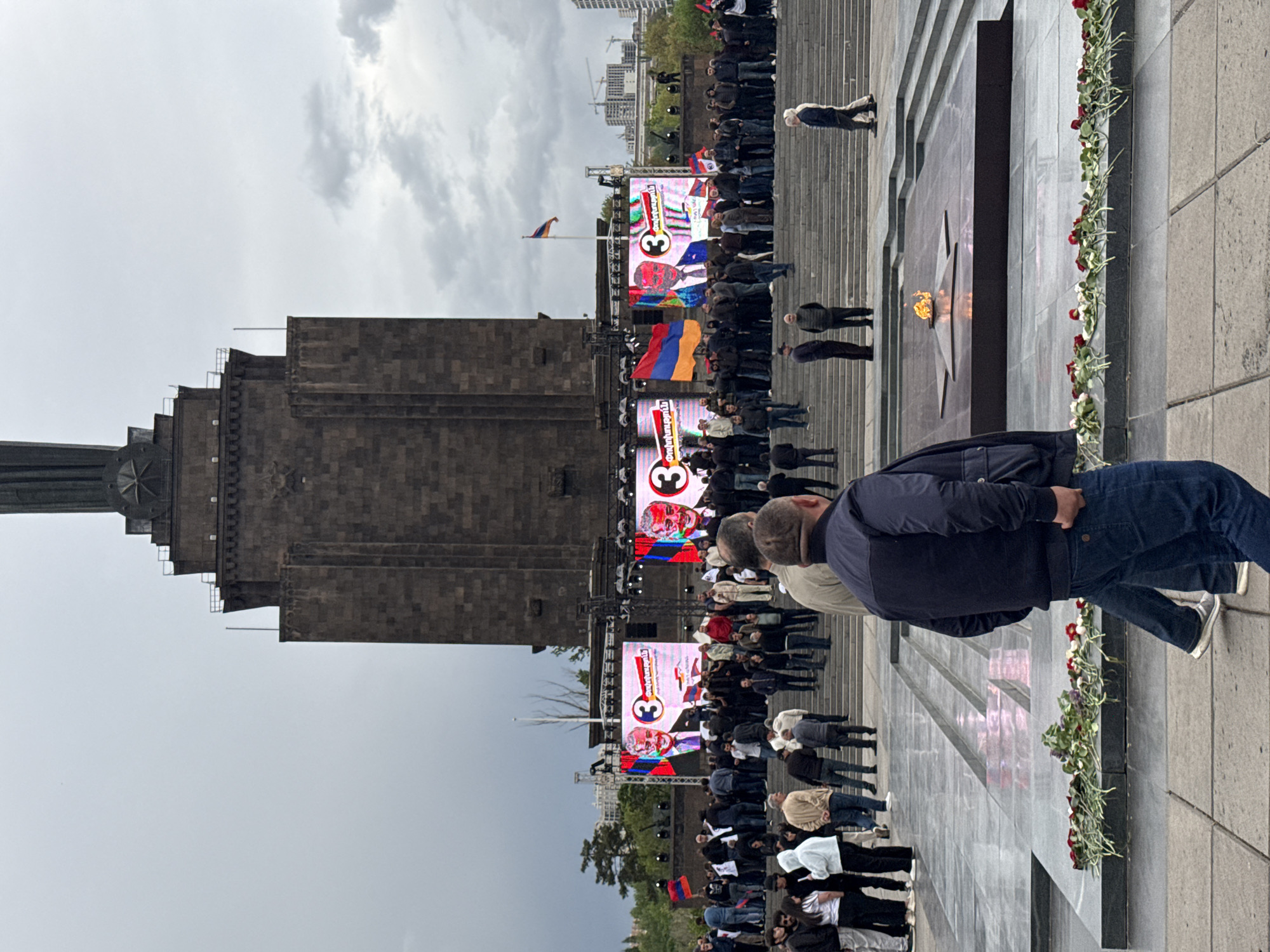 The image shows the Yerevan Genocide Memorial Complex, situated in Yerevan, Armenia. The memorial is a large stone structure. In front of the structure is a public gathering of people, possibly an event. Screens displaying images and text are visible, along with the Armenian flag. A flame burns within a central star-shaped monument. Flowers are laid along the edges. The sky is overcast. People are standing and observing. Modern buildings are visible in the background.