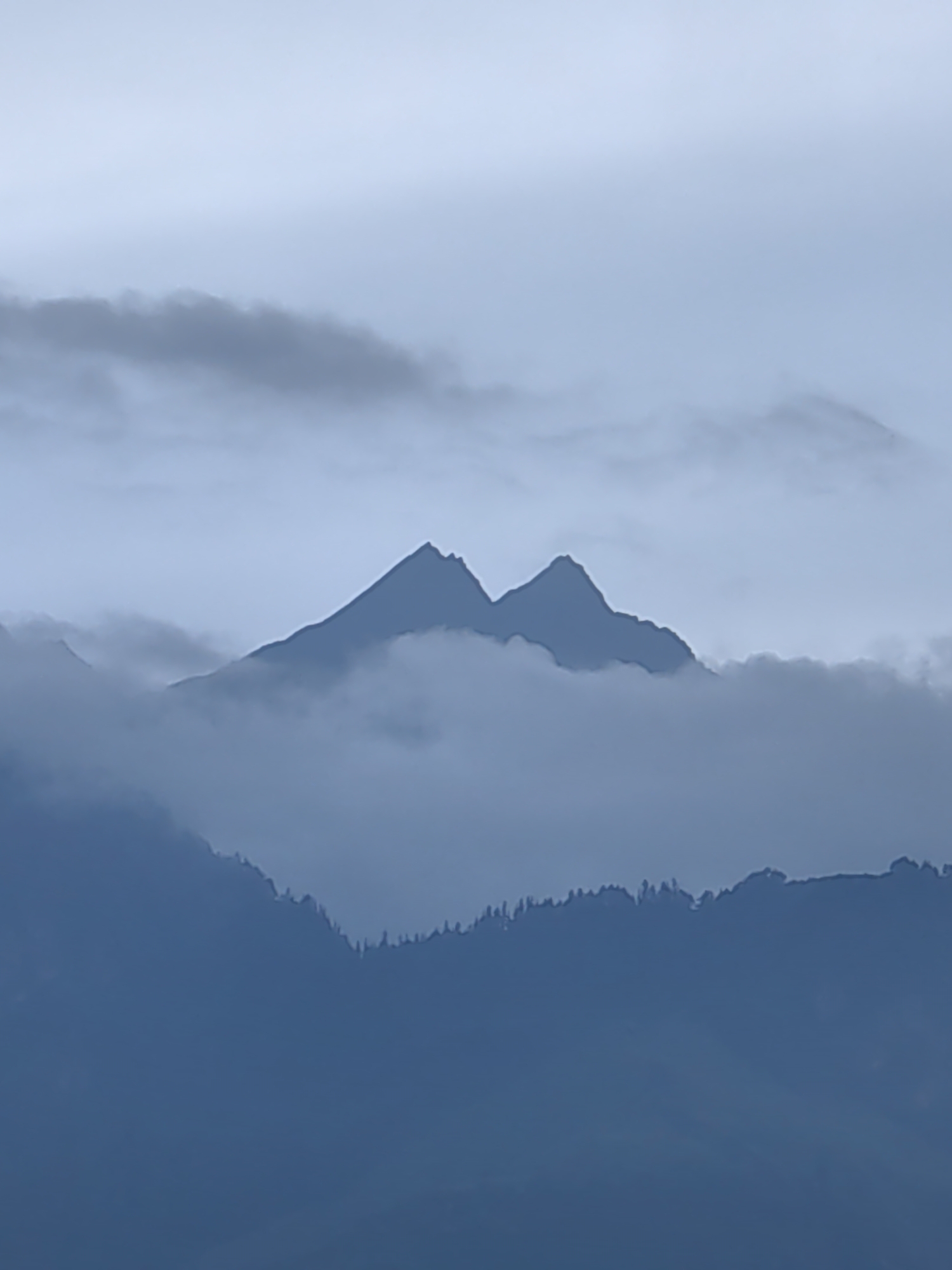 The image shows a pair of jagged, snow-capped mountain peaks emerging from a sea of clouds. The mountains are silhouetted against a pale, overcast sky. In the foreground, darker, lower mountains are visible, their slopes covered in a dense, dark green forest. The overall mood is one of dramatic, ethereal beauty, with the mist and clouds obscuring the base of the mountains and giving them a mystical appearance. The scene suggests a remote, high-altitude location.