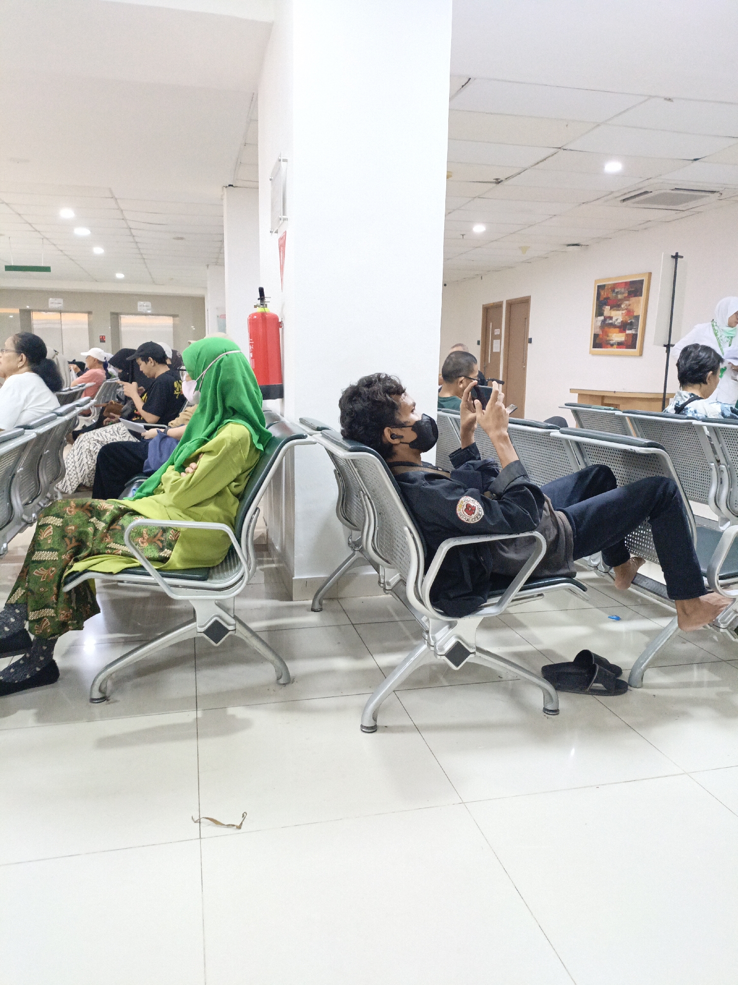 The image depicts a waiting area, likely in a hospital or clinic, within East Jakarta, Indonesia. The scene is characterized by rows of metal chairs, typical of public waiting spaces. Several individuals are seated, with varying postures and engagement. In the foreground, a person wearing a black shirt and mask is engrossed in their mobile phone, seated with their legs extended. To the left, a woman in a lime green hijab and patterned batik skirt is also seated, her head bowed. Other individuals are visible in the background, some looking at their phones, others seemingly in conversation or resting. A bright red fire extinguisher is mounted on a white pillar, a common safety feature in such environments. The floor is tiled in a light color, and overhead fluorescent lights illuminate the space. A framed artwork hangs on a distant wall, adding a touch of decor. The overall impression is one of quiet anticipation, with individuals waiting in a public, indoor setting.