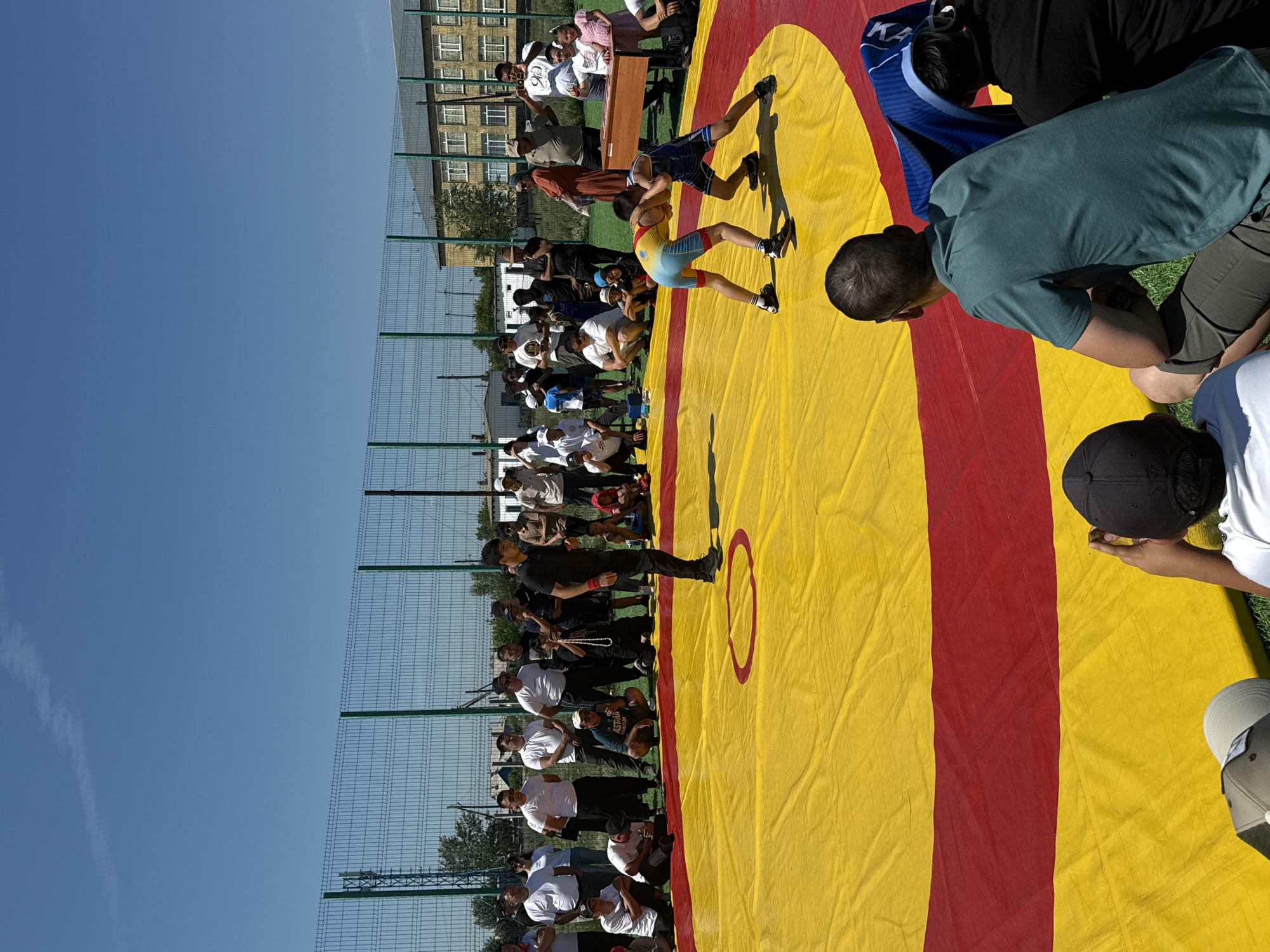 In the city of Өндіріс, Kazakhstan, an outdoor wrestling match is underway on a bright, sunny day. A vibrant yellow wrestling mat with a red border serves as the arena, with a smaller red circle marking the center. Two young wrestlers, one in a yellow and blue singlet and the other in a blue singlet, are engaged in a grapple. A referee, dressed in black, stands nearby, observing the match. A crowd of spectators, dressed in casual attire, surrounds the mat, some seated on the grass and others standing. In the background, a tall chain-link fence encloses the area, with buildings and trees visible beyond. The sky is clear and blue, indicating pleasant weather conditions for the event.