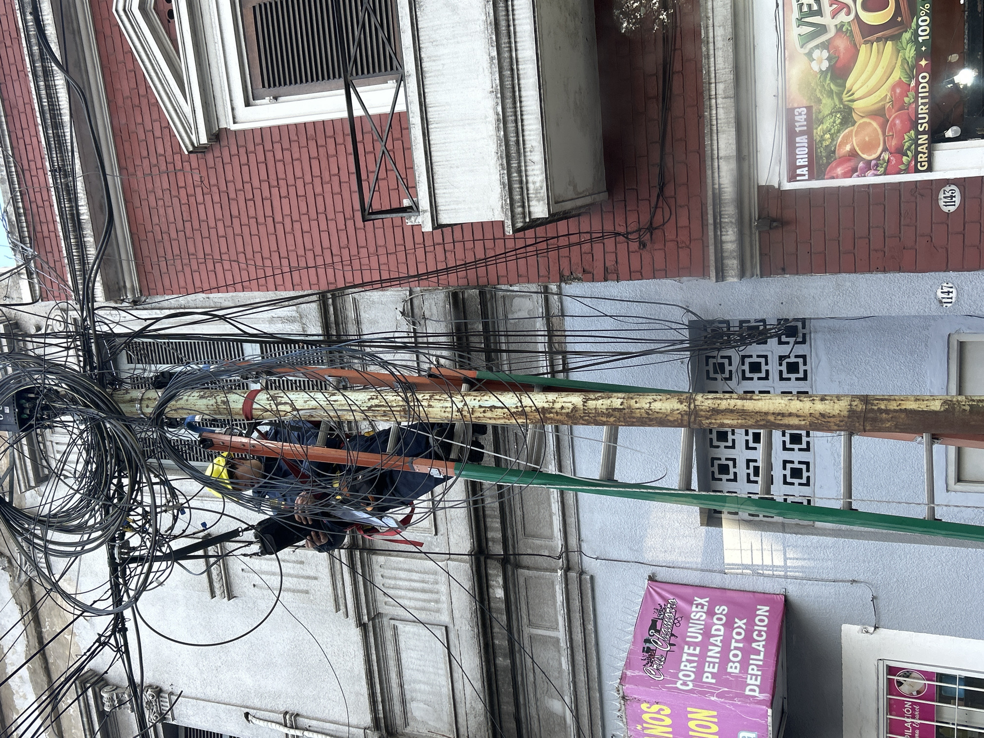 A person wearing a yellow hard hat and a dark blue uniform is working on a utility pole tangled with numerous electrical wires. The person is positioned on a ladder, which appears to be leaning against the pole, and is holding what looks like a tool or equipment in their hands. The utility pole is metallic and shows signs of rust. Behind the pole, there's a light blue building with some decorative architectural elements. To the right of the pole is a red brick building with a balcony. A sign on the blue building, partially visible, appears to be for a hair salon, listing services like "CORTE UNISEX," "PEINADOS," "BOTAX," and "DEPILACION." On the red brick building, a sign advertises "LA RIOJA 1143" and features images of fruits and vegetables. The overall scene suggests an urban environment in Buenos Aires, Argentina, with a focus on utility work and commercial activity. The presence of the street numbers "1147" and "1143" on the buildings further supports this location.