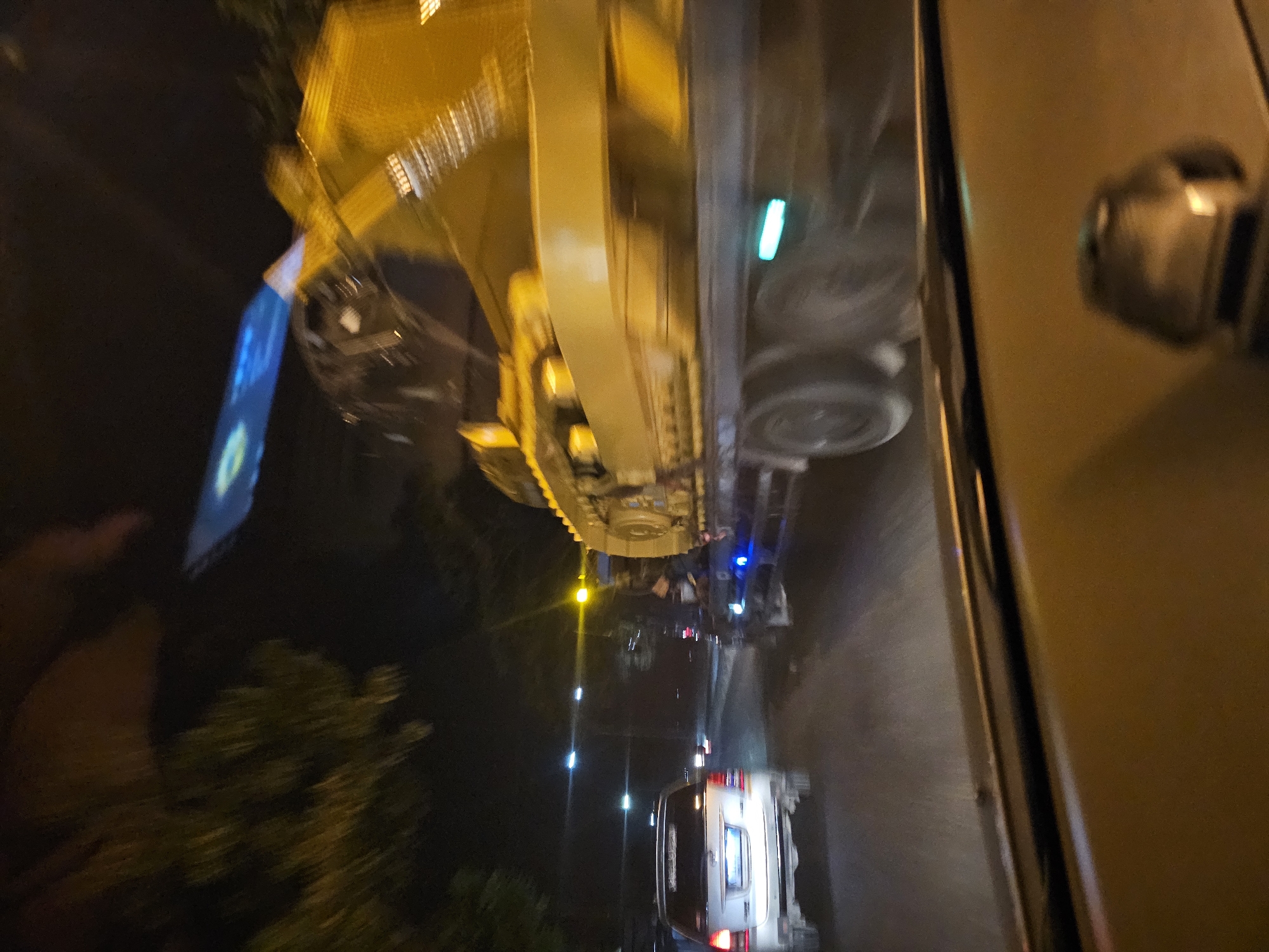 In Waruwetan, Indonesia, at night, a silver SUV is traveling on a road. A yellow excavator is being transported on a flatbed truck, also on the road. The scene is dimly lit by streetlights and vehicle headlights. The image appears to be taken from inside another vehicle, possibly the SUV, due to the perspective and the view of the dashboard. The overall impression is one of nighttime traffic on a city road in Indonesia.