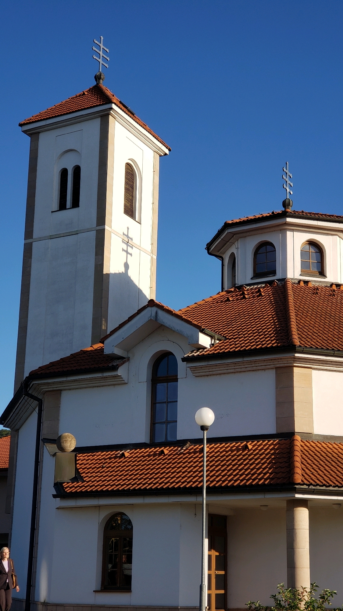 The image shows a close-up, low-angle view of a modern church building against a clear blue sky. The church features white walls, a prominent bell tower with a cross on top, and a domed section also topped with a cross. The roofs are covered in reddish-brown tiles. A shadow of a cross is cast on the side of the bell tower. A street lamp with a spherical light fixture stands in front of the building. To the left, a woman in dark clothing walks away from the camera. The architecture suggests a location in Eastern Europe, possibly Prešov, Slovakia, given the style of the crosses and the building. The scene is bathed in sunlight, creating distinct shadows.